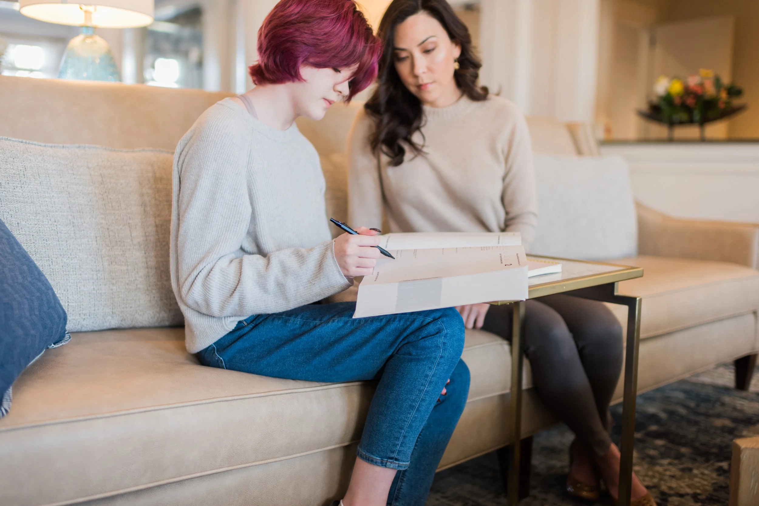 Two women sitting on a beige sofa in a cozy, well-lit living room, looking at a book or notebook together. One woman has short, wavy pink hair and is wearing a light-colored sweater and jeans. The other woman has long, dark hair, wearing a cream-colored sweater, and is looking at the book with interest. A table with a floral arrangement is visible in the background.