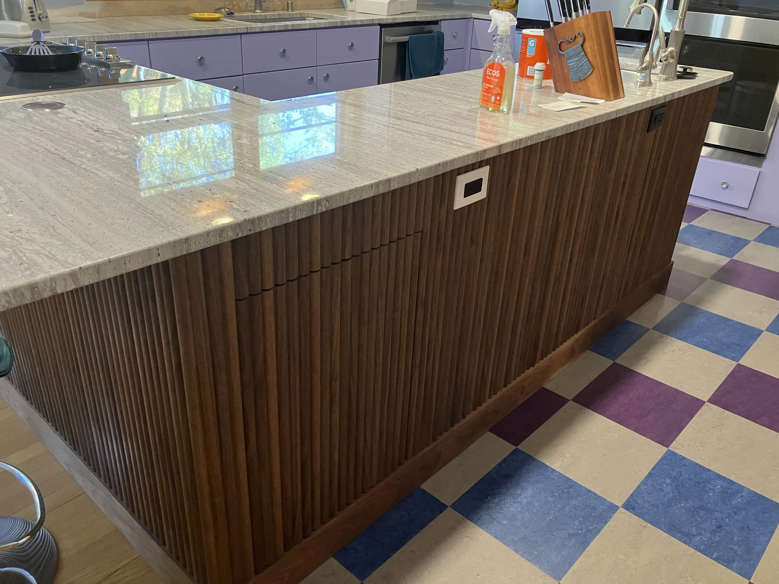 Kitchen island with a marble countertop, wooden paneling on the sides, and various cleaning supplies and paper products on top. The floor has a colorful checkered pattern with purple, blue, and beige tiles.