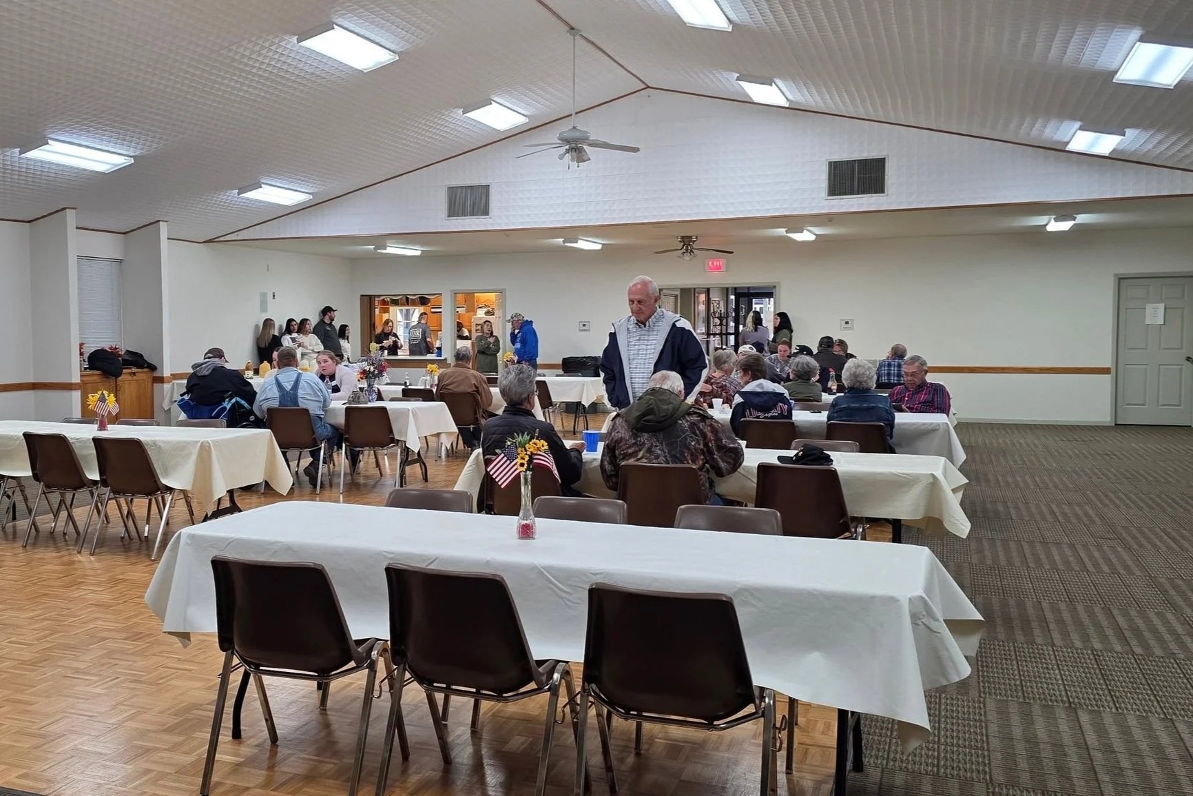 Community hall with people sitting at tables decorated with small American flags and patriotic flowers, some standing and chatting, suggesting a patriotic or holiday gathering.