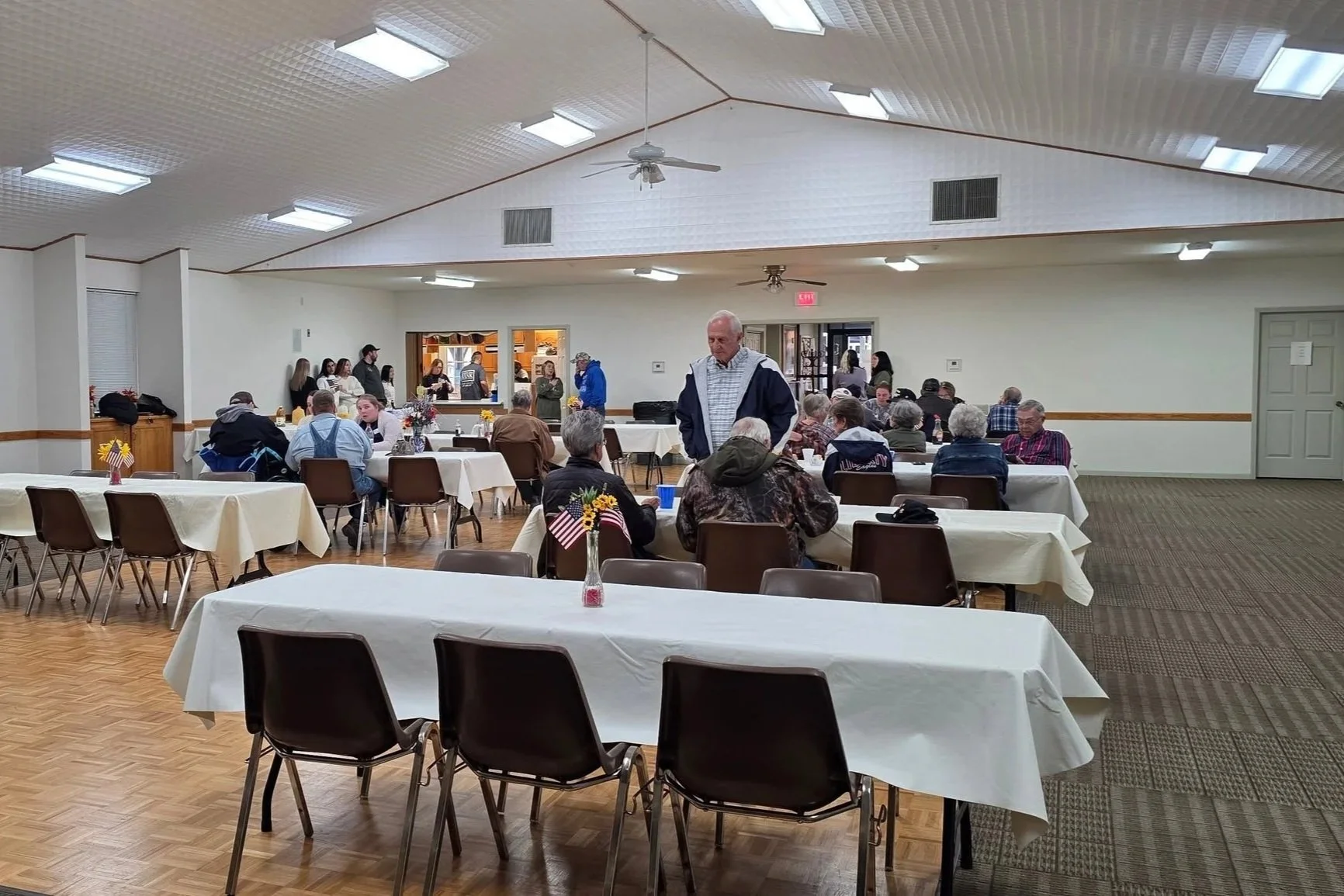 A community gathering in a hall decorated with American flags and flowers; people sitting at tables covered with white tablecloths, some conversing, others waiting; a counter area in the background with a few people behind it; the hall has a high ceiling with white paneling and fluorescent lighting.
