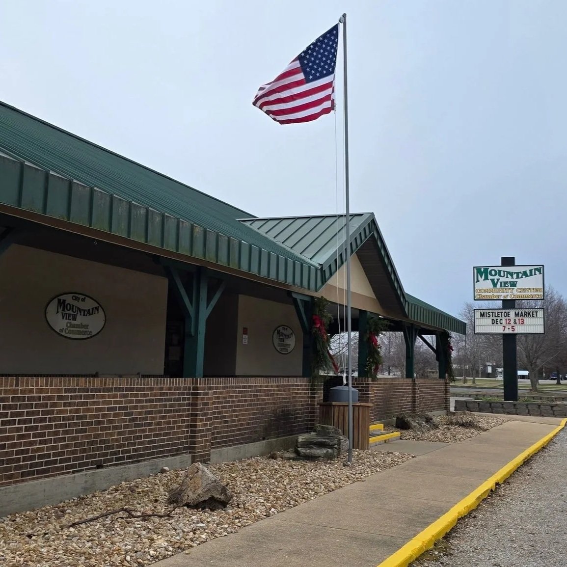 American flag flying outside of a brick building with a green metal roof, with a sign in the background that reads 'Mountain View Chamber of Commerce' and advertising upcoming holiday events.
