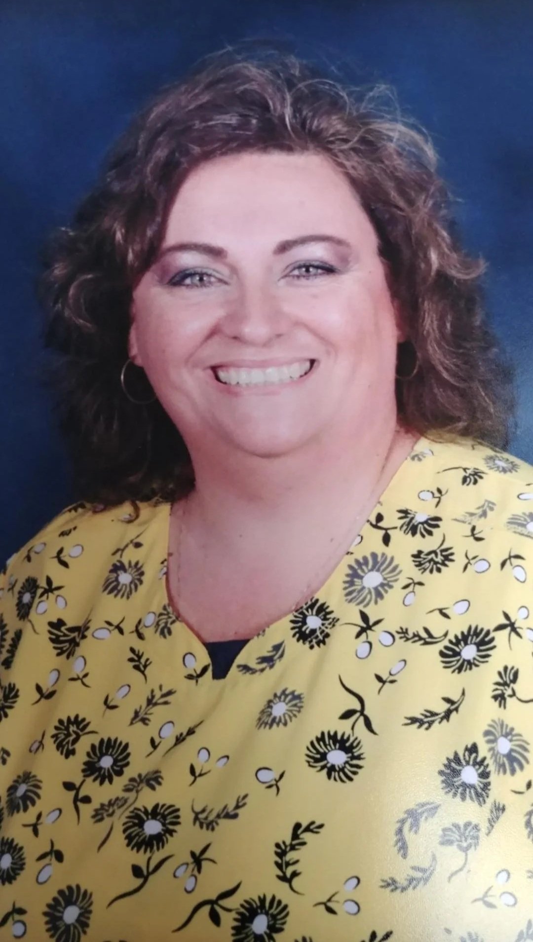 A woman with short curly brown hair, wearing a yellow top with a floral pattern and hoop earrings, smiling against a dark blue background.