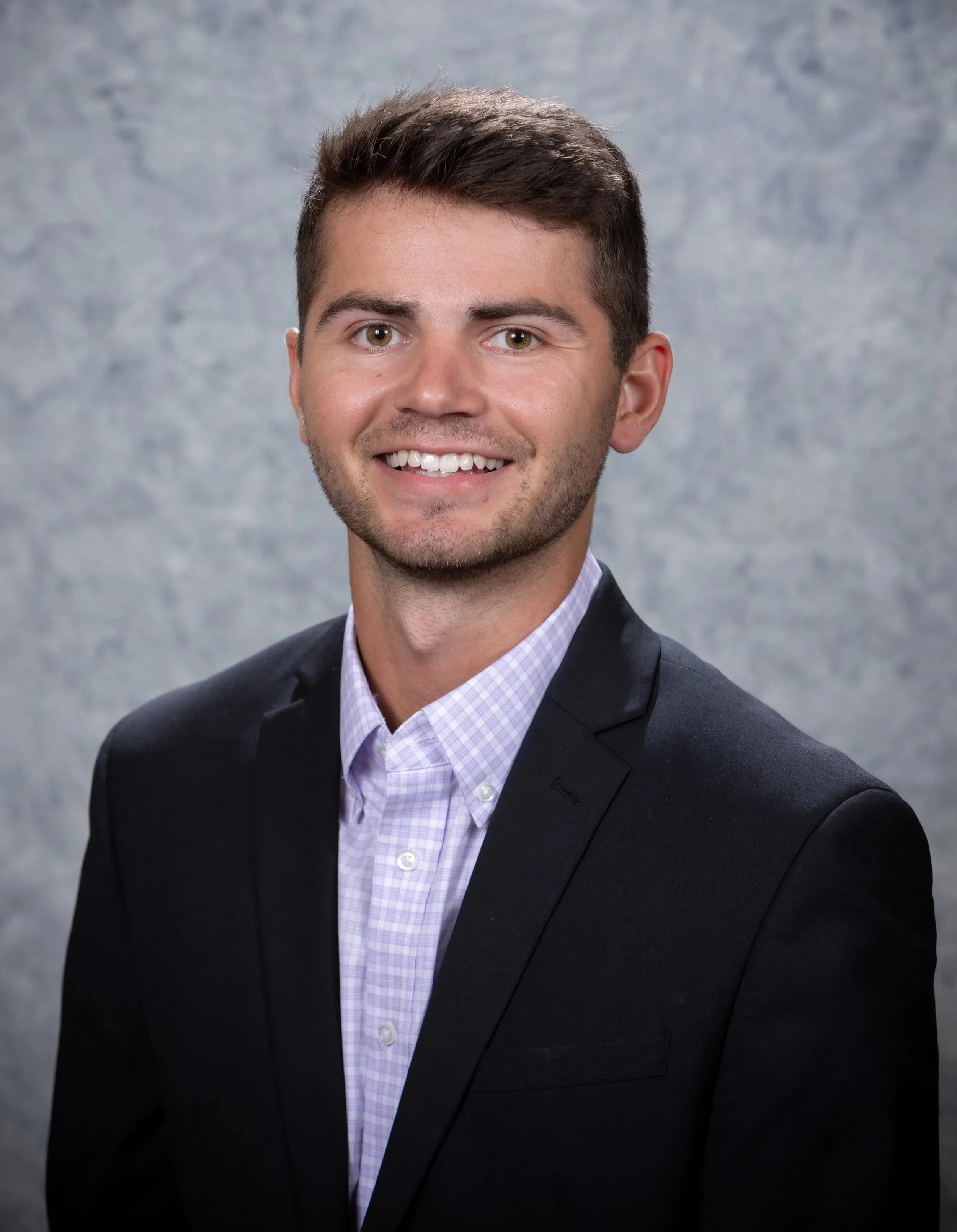 Portrait of a young man smiling, standing in front of a wall with colorful abstract paintings, wearing a gray zip-up vest with 'BLUESTONE FINANCIAL ADVISORS' logo.