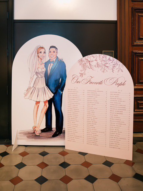 Vogue Vignette life-size custom illustrated bride and groom selfie wall cutout displayed alongside a bespoke illustrated seating chart at a wedding reception, photographed by May Carlson.