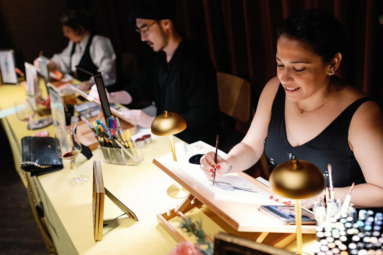 Vogue Vignette artists creating live guest portraits at a wedding reception, sketching by hand at an illuminated on-site illustration studio.