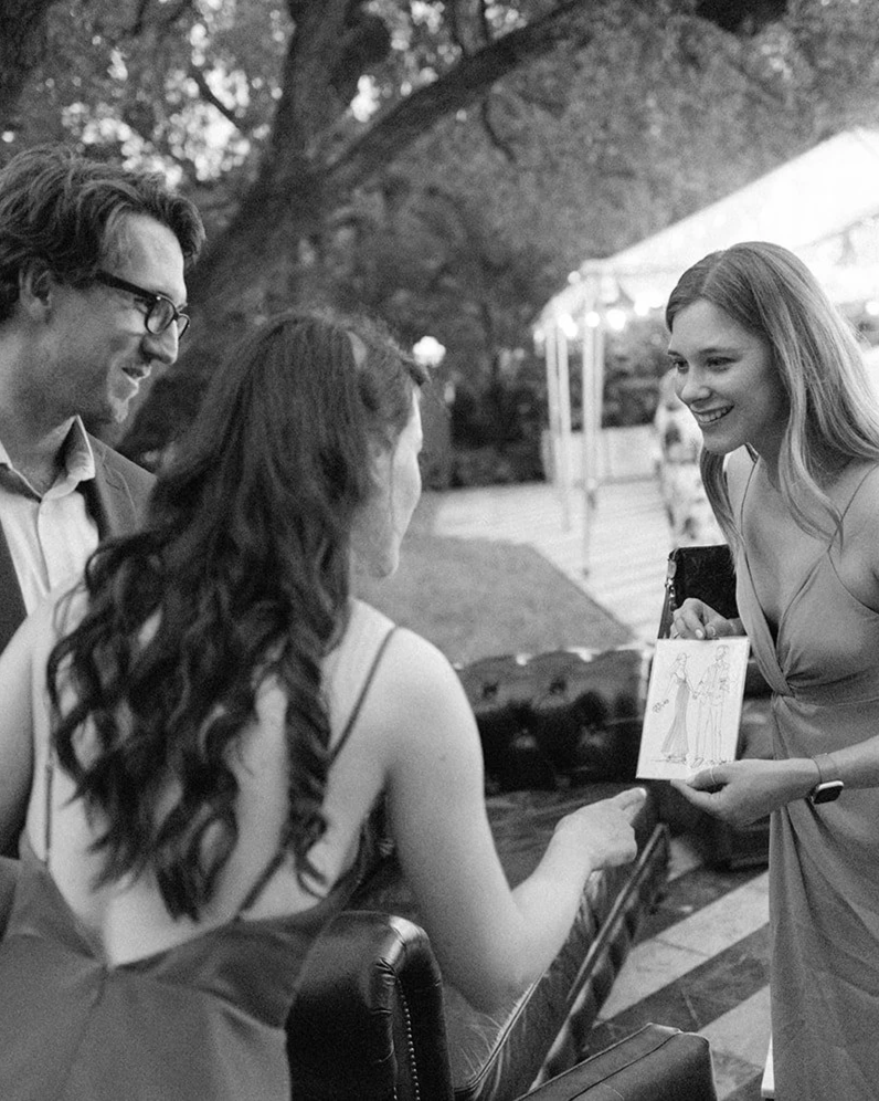 Three people at an outdoor wedding: a man with glasses and a woman with curly hair are smiling at a woman with long straight hair who is holding a drawing and a photo.
