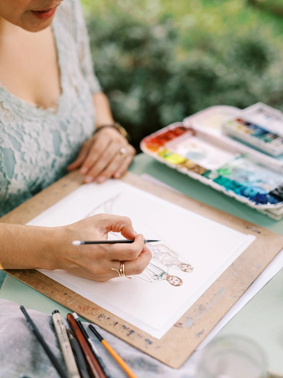 Vogue Vignette artist Stephanie Schiller sketching a live guest portrait by hand at a wedding, with watercolor paints and fine art pens on an outdoor illustration setup.