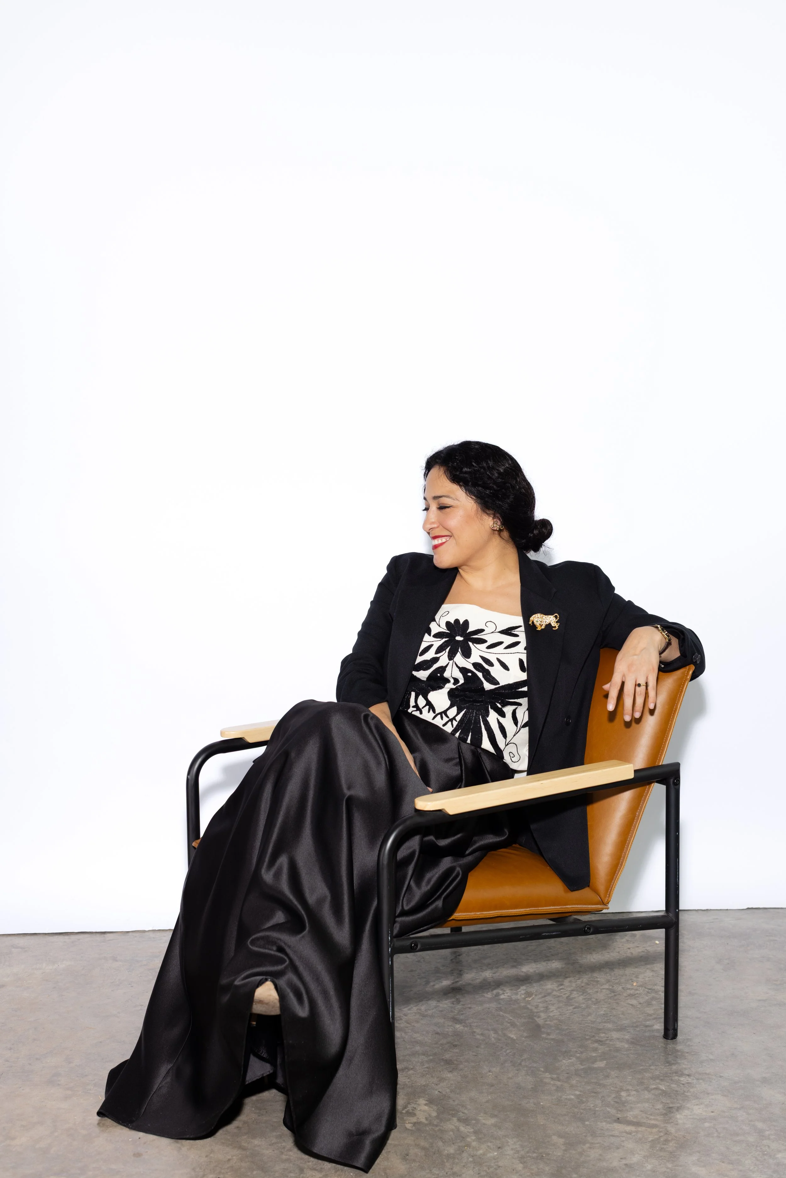 Woman in a black blazer and patterned blouse, Stephanie J. Schiller, smiling and sitting on a brown chair with black metal frame, in front of a white wall.