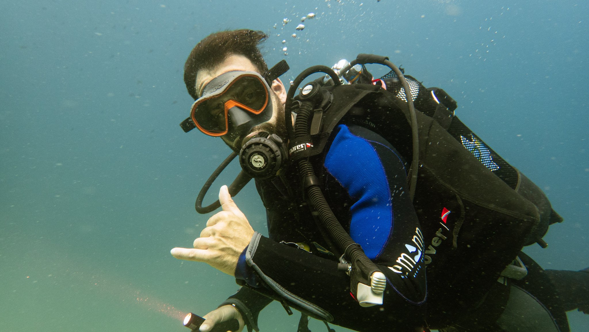 A man scuba diving underwater wearing a black wetsuit with blue accents, orange and black diving mask, and breathing through a regulator. He is giving a shaka sign with his hand.