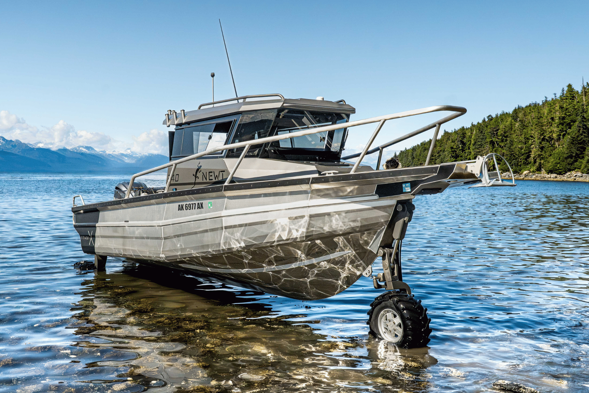 A modern 240UCC StabiX amphibious boat in Alaska, coming out of the water with snow-capped mountains and green forested hills in the background.