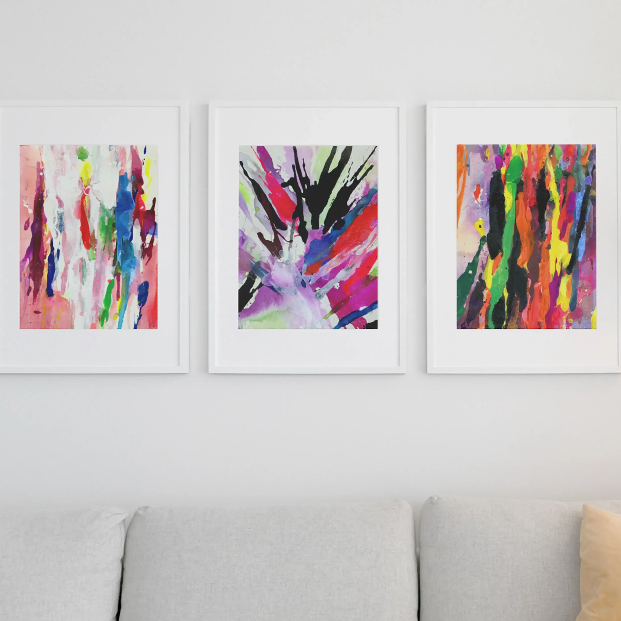 Three framed abstract watercolor paintings featuring pastel drips, a bold black burst, and vibrant vertical strokes displayed above a light gray sofa on a white wall in a modern living room.