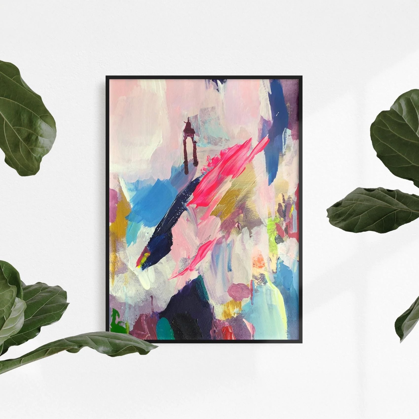 Colorful abstract artwork framed in black and hung on a white wall surrounded by large green leaves, creating a bright, fresh interior decor setting.