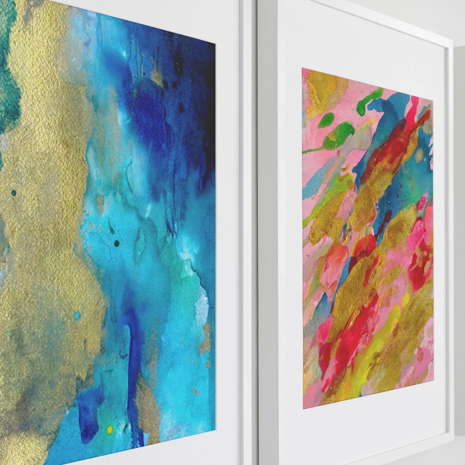 Side view of two framed abstract paintings featuring vibrant blue and pink color splashes with gold highlights, hung in a modern gallery-style room