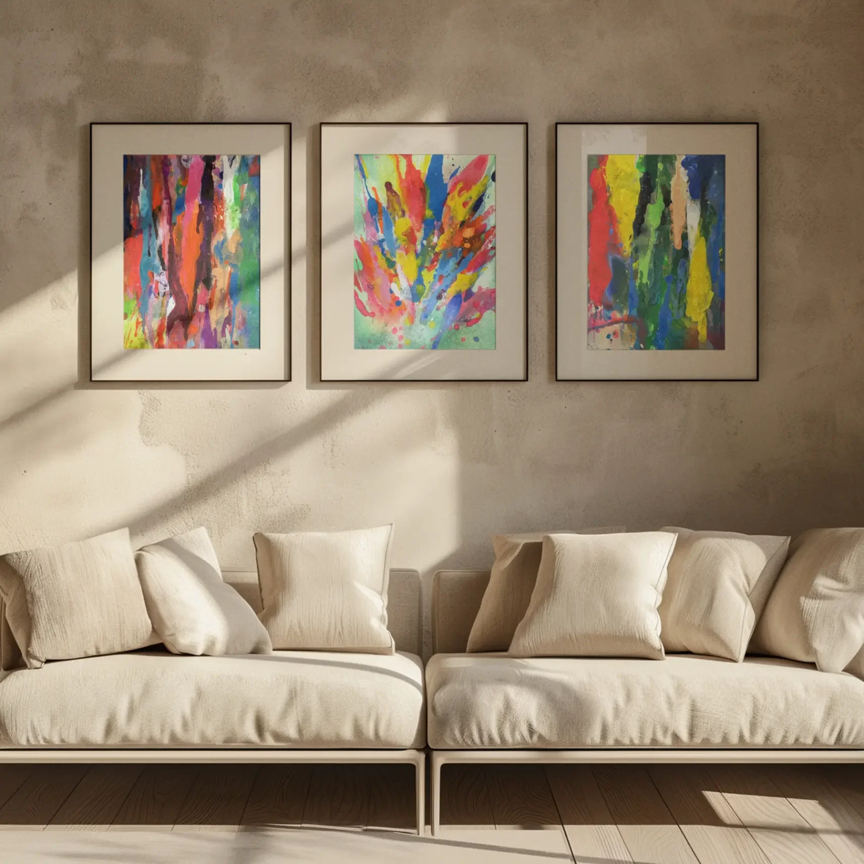 Three framed abstract watercolor paintings with vibrant multicolor drips and splashes displayed above a neutral beige sofa in a modern living room with warm natural light.