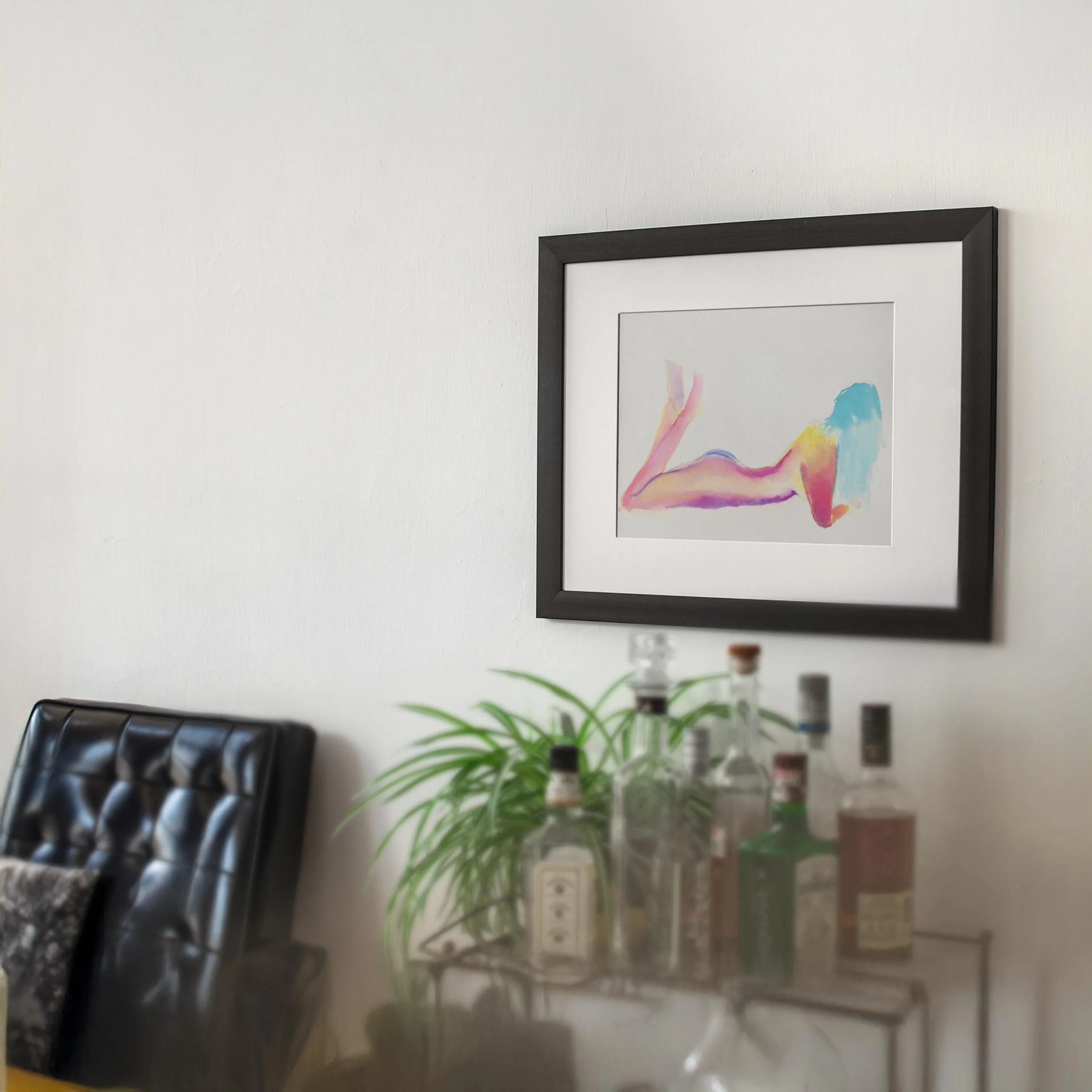 Vacation_8.jpg Framed watercolor nude in neon pink, orange, and cyan tones, hanging above a bar cart in a cozy home interior with a black leather chair and houseplant.