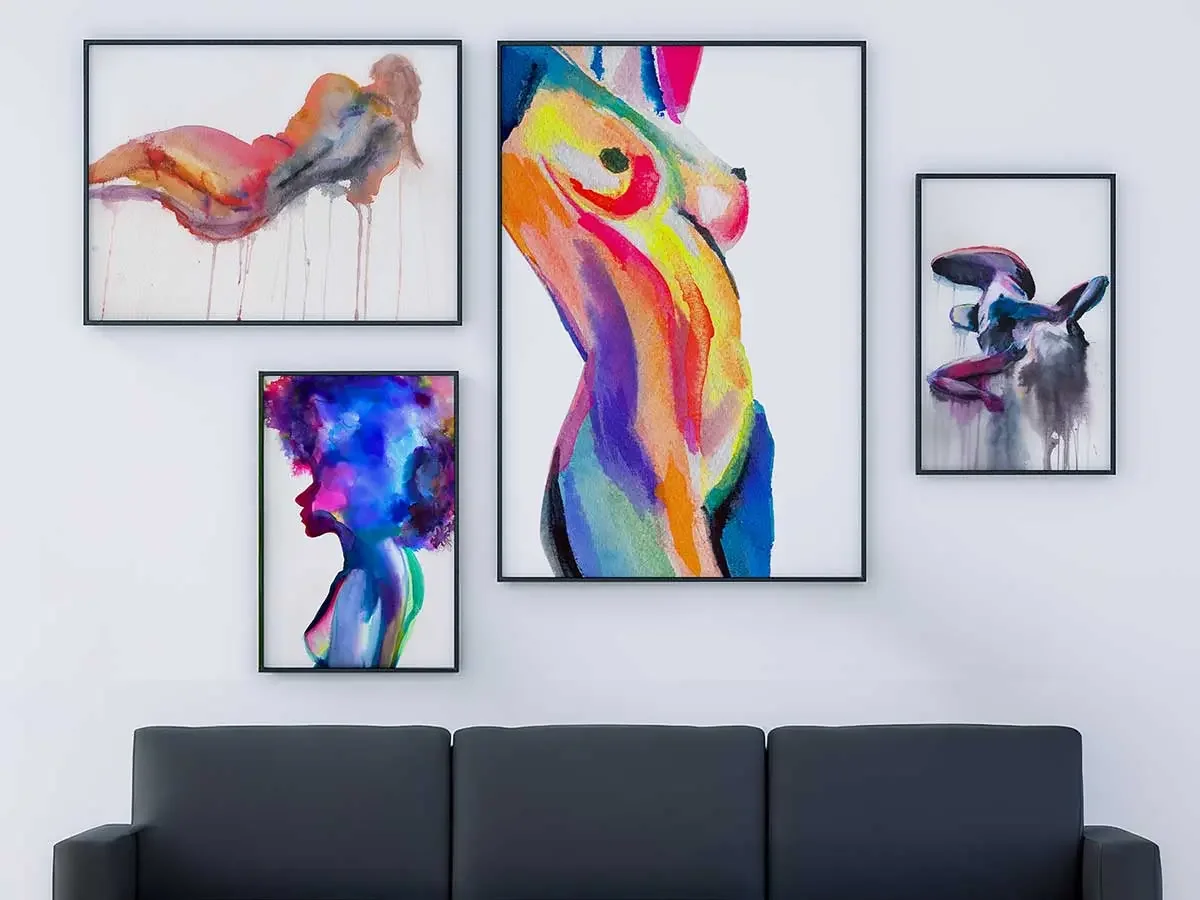 Gallery wall display of colorful abstract nude watercolor original paintings in black frames above a modern gray sofa, featuring vibrant expressive female figures in pink, blue, yellow, and purple tones.