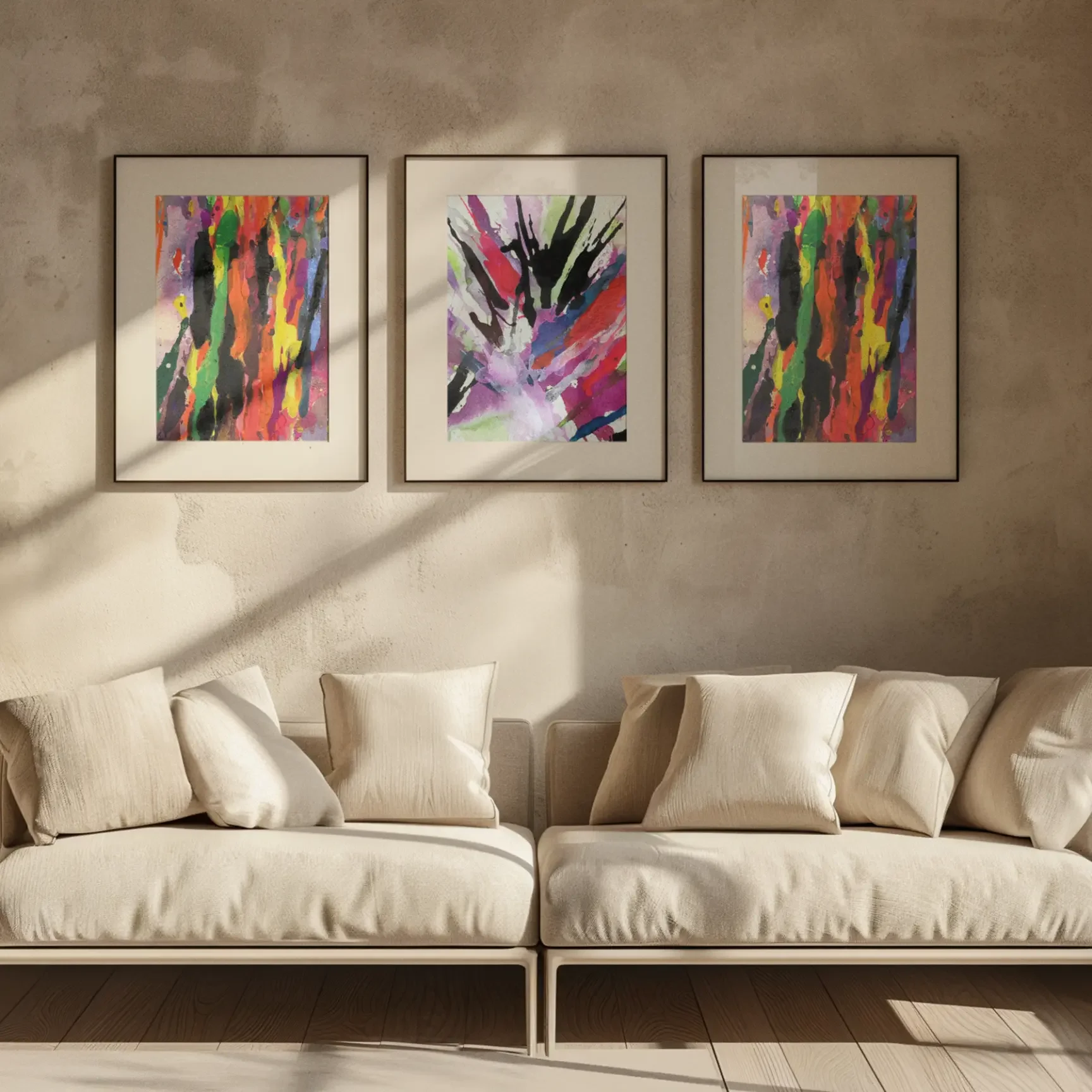 Three framed abstract watercolor paintings with bold multicolor drips and a central burst design displayed above a neutral beige sofa in a modern living room with warm natural light.