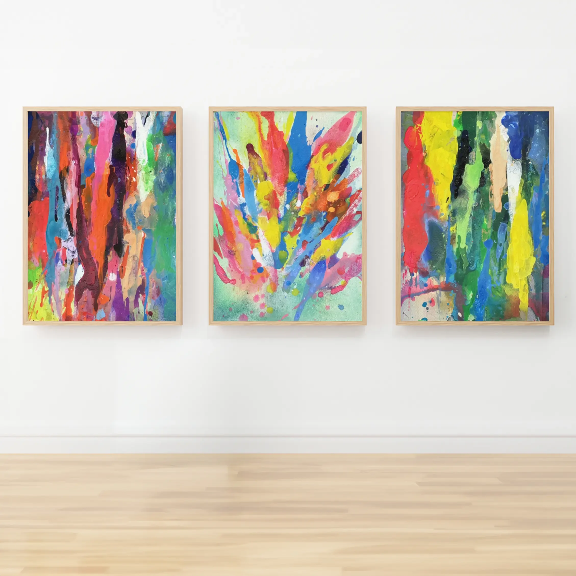 Three colorful abstract watercolor paintings with bold drips and splashes in red, yellow, blue, green, and pink, displayed side by side in natural wood frames on a white wall above a light wood floor.