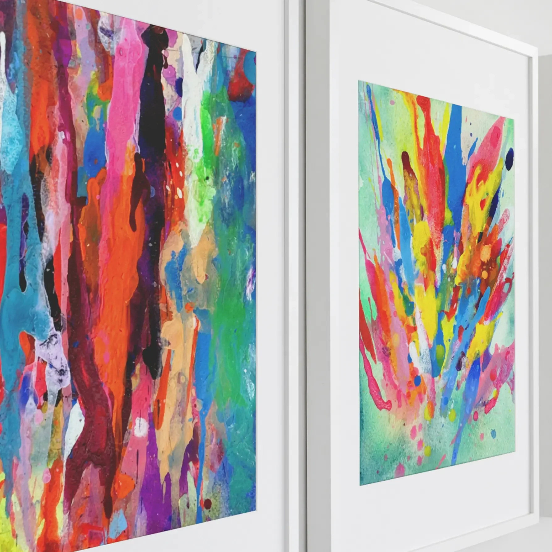 Close-up of two framed abstract watercolor paintings with vibrant multicolor drips and splashes in pink, blue, green, orange, and yellow, displayed side by side in white frames against a neutral wall.