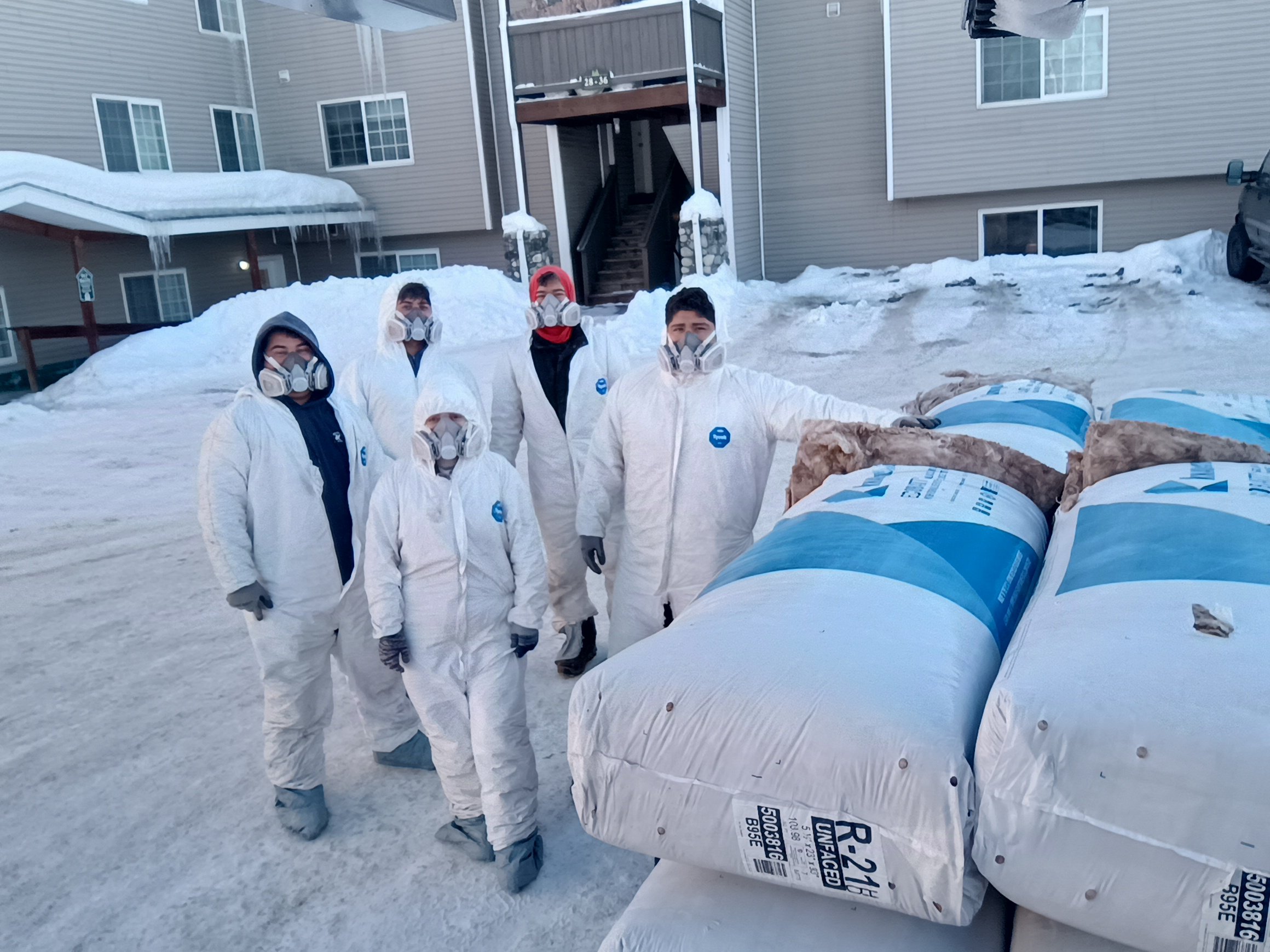 People wearing protective suits and respirators standing outdoors in a snowy environment near large bags labeled R-21 unfaced insulation.