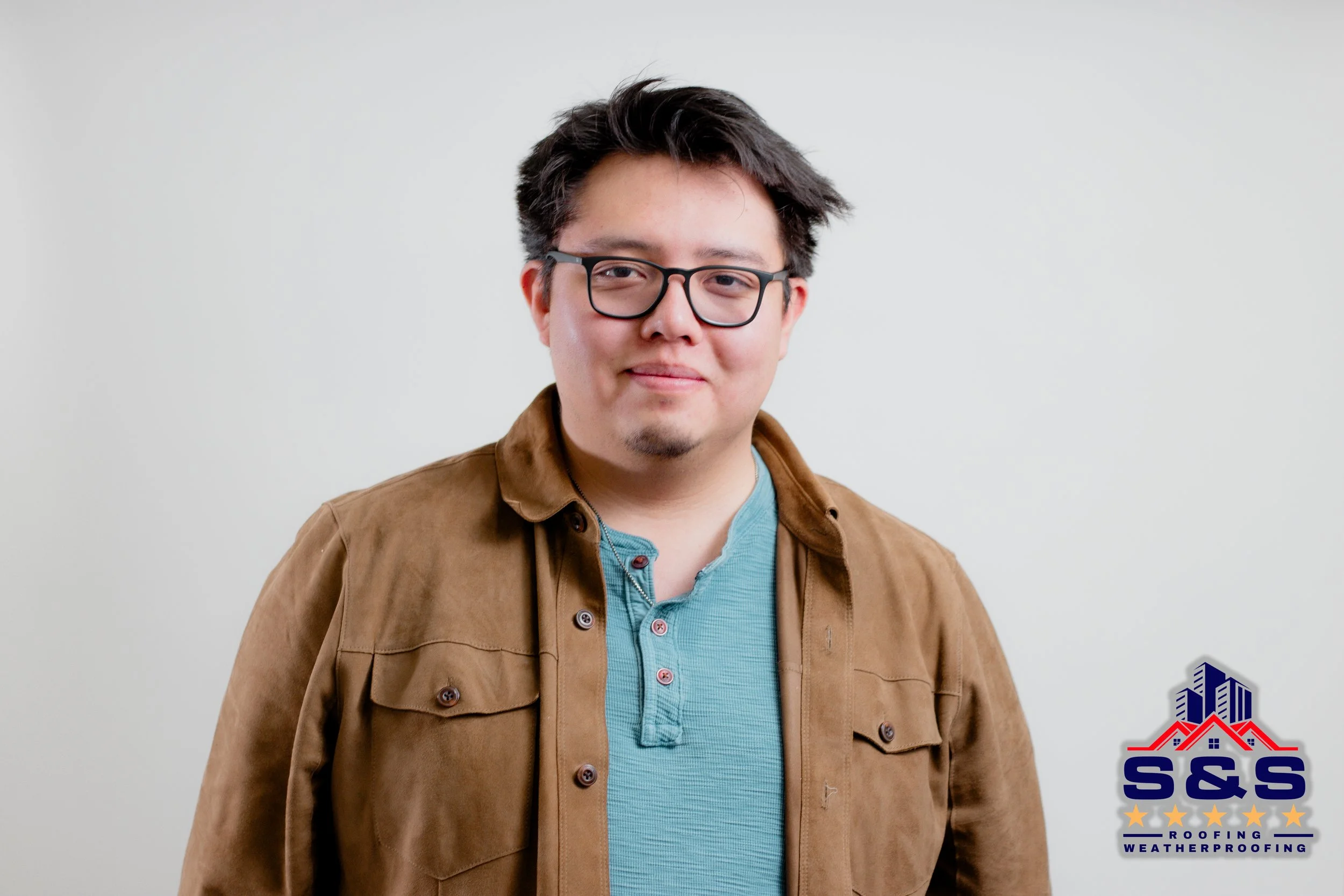 A smiling young man with arms crossed, wearing a gray collared shirt with a company logo, standing in front of a blue pickup truck on a cloudy day.