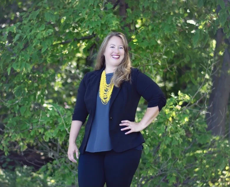 A woman with long blonde hair smiling outdoors, wearing a black blazer, dark blouse, black pants, and a yellow necklace, standing in front of green trees.