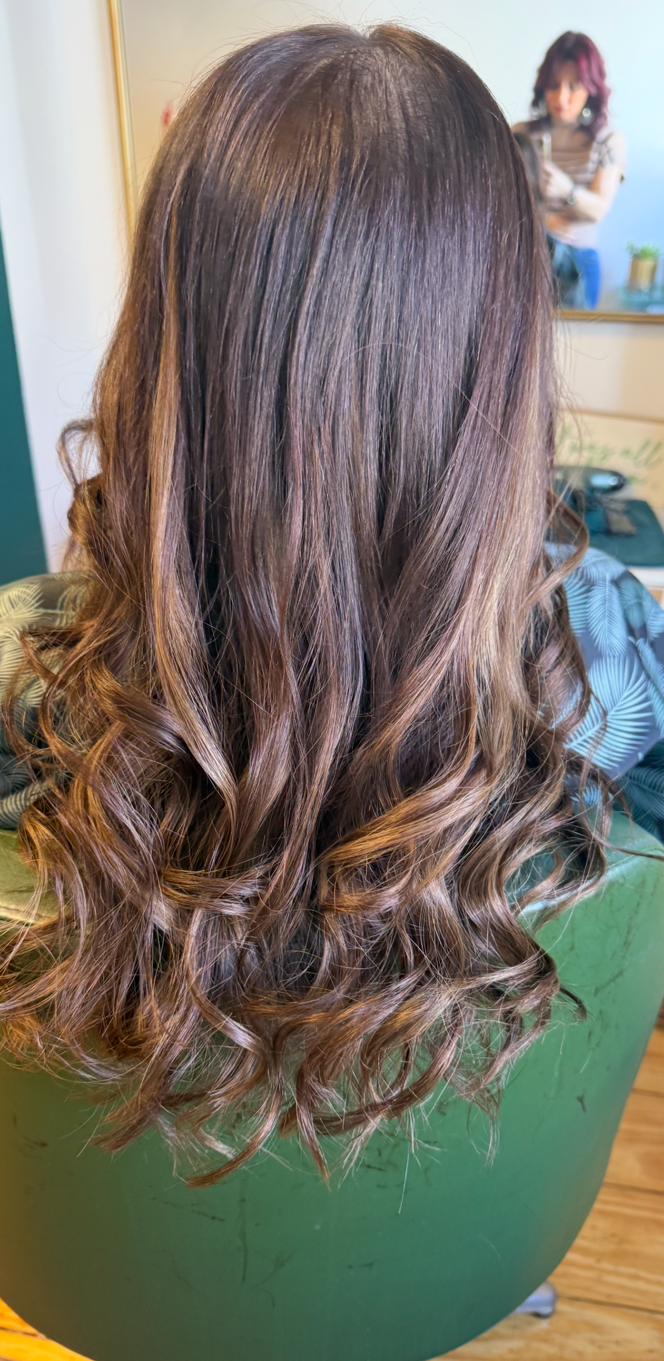 Brunette woman with long, wavy hair sitting in a green salon chair, with another woman in the background taking a photo