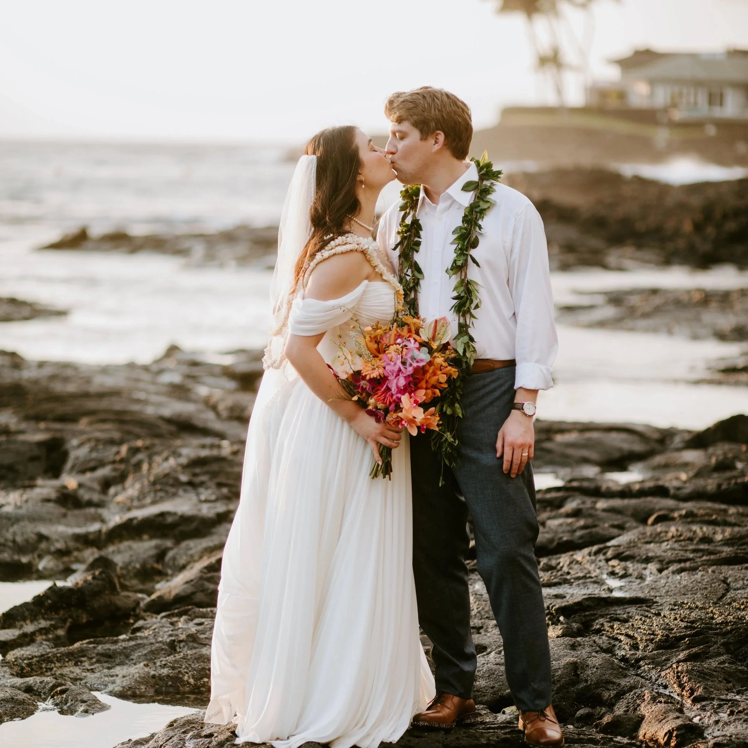 Bride and groom sharing a kiss on a rocky beach at sunset, with the bride holding a colorful bouquet, wearing flowy white dress, and the groom in a white shirt and dark pants, both adorned with a green leaf lei.