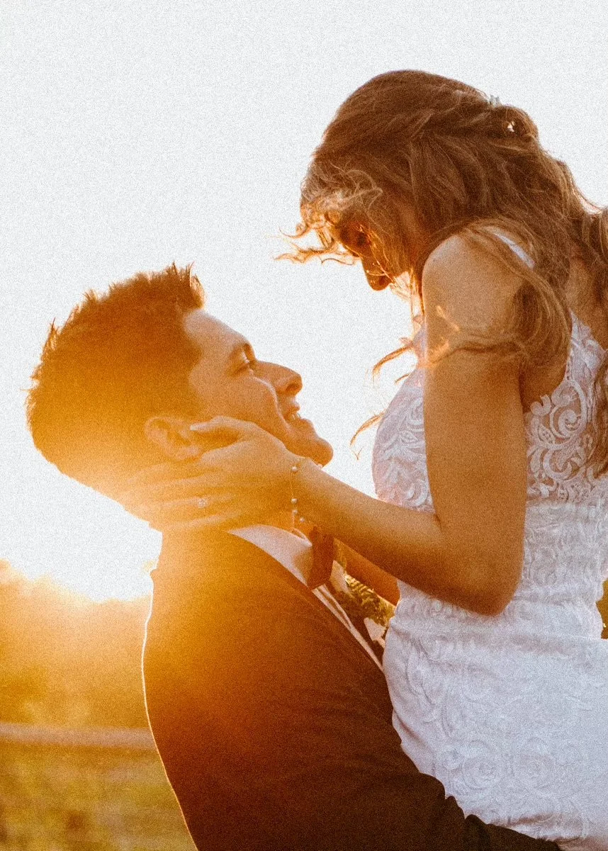 A man lifting a woman in a white dress during sunset, both smiling and gazing at each other lovingly.