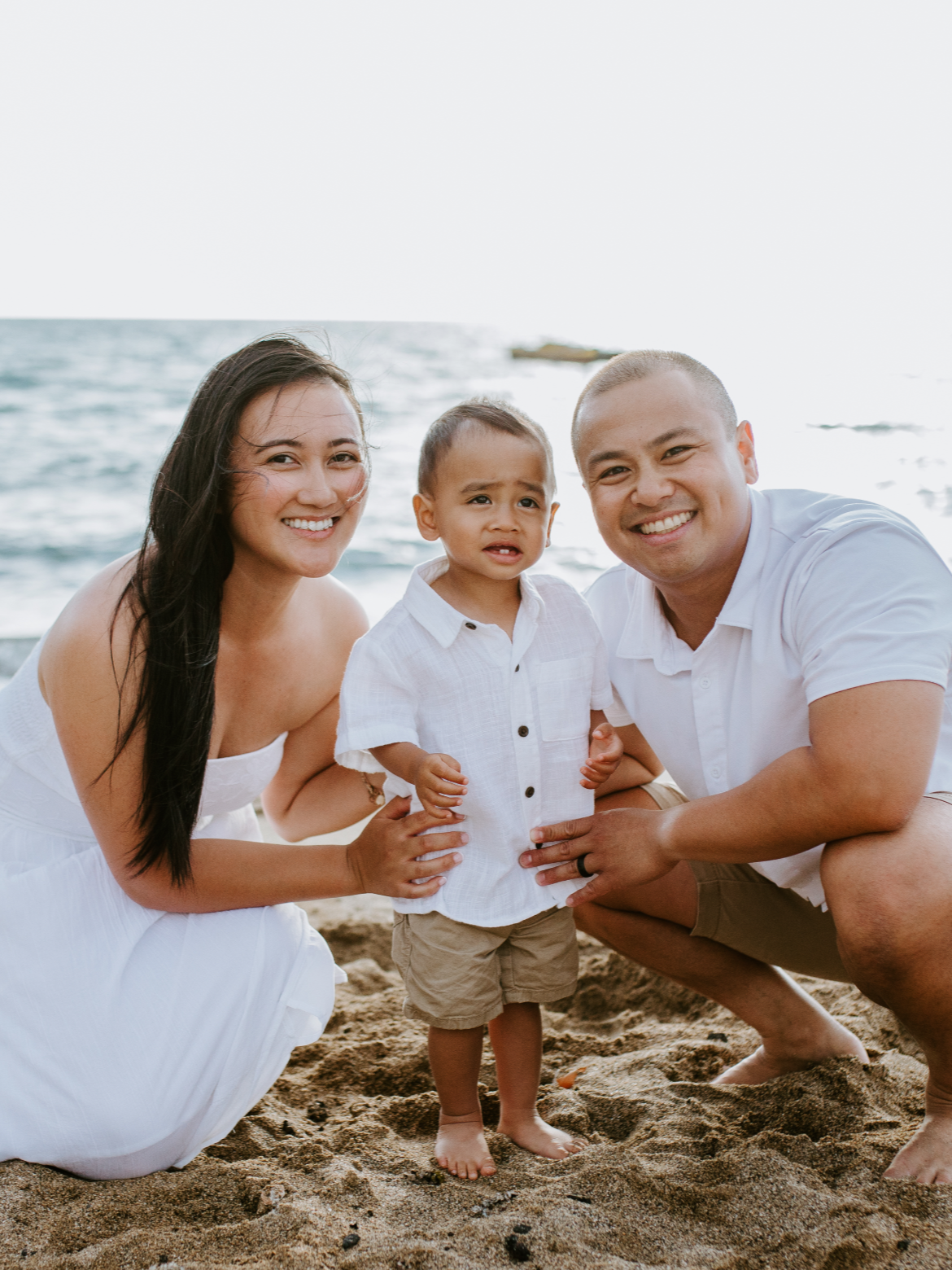 A happy family of three, including a woman, a man, and a young boy, poses on the beach with the ocean in the background, during daytime.