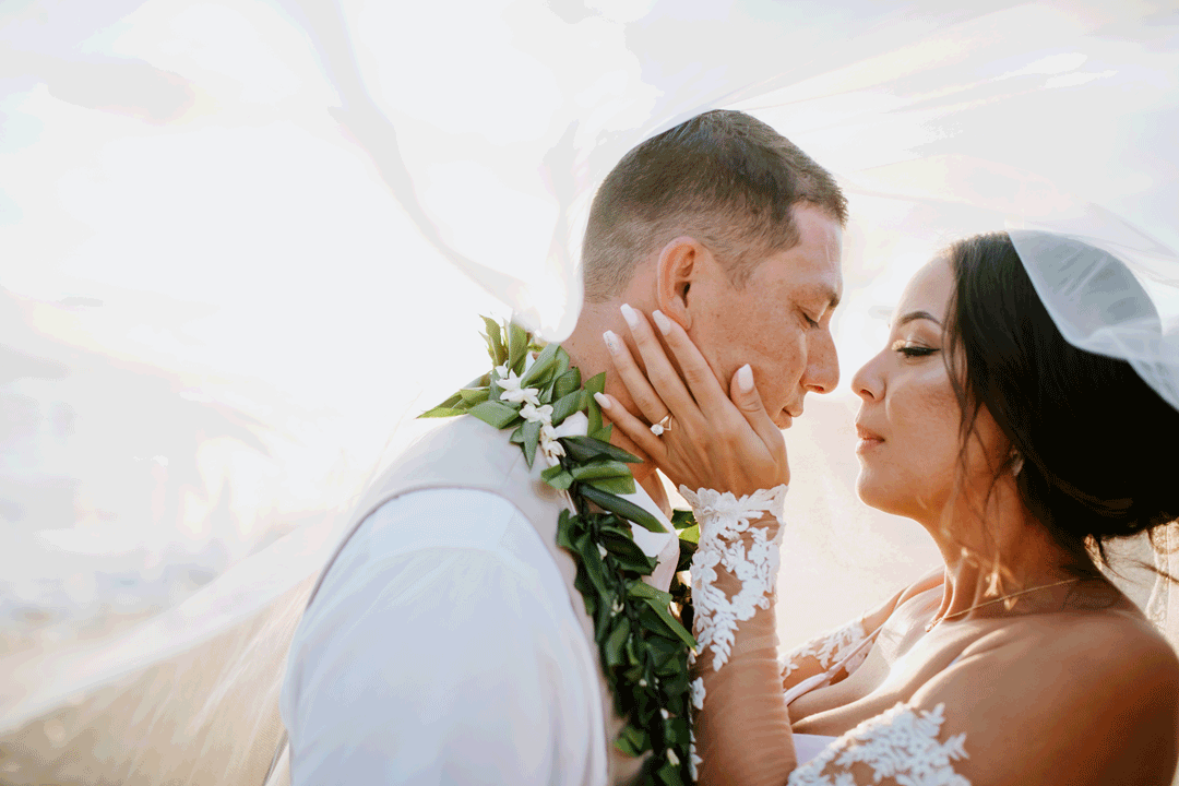 A bride and groom share an intimate moment outdoors, with the bride gently touching the groom's face under his chin, both with their eyes closed.