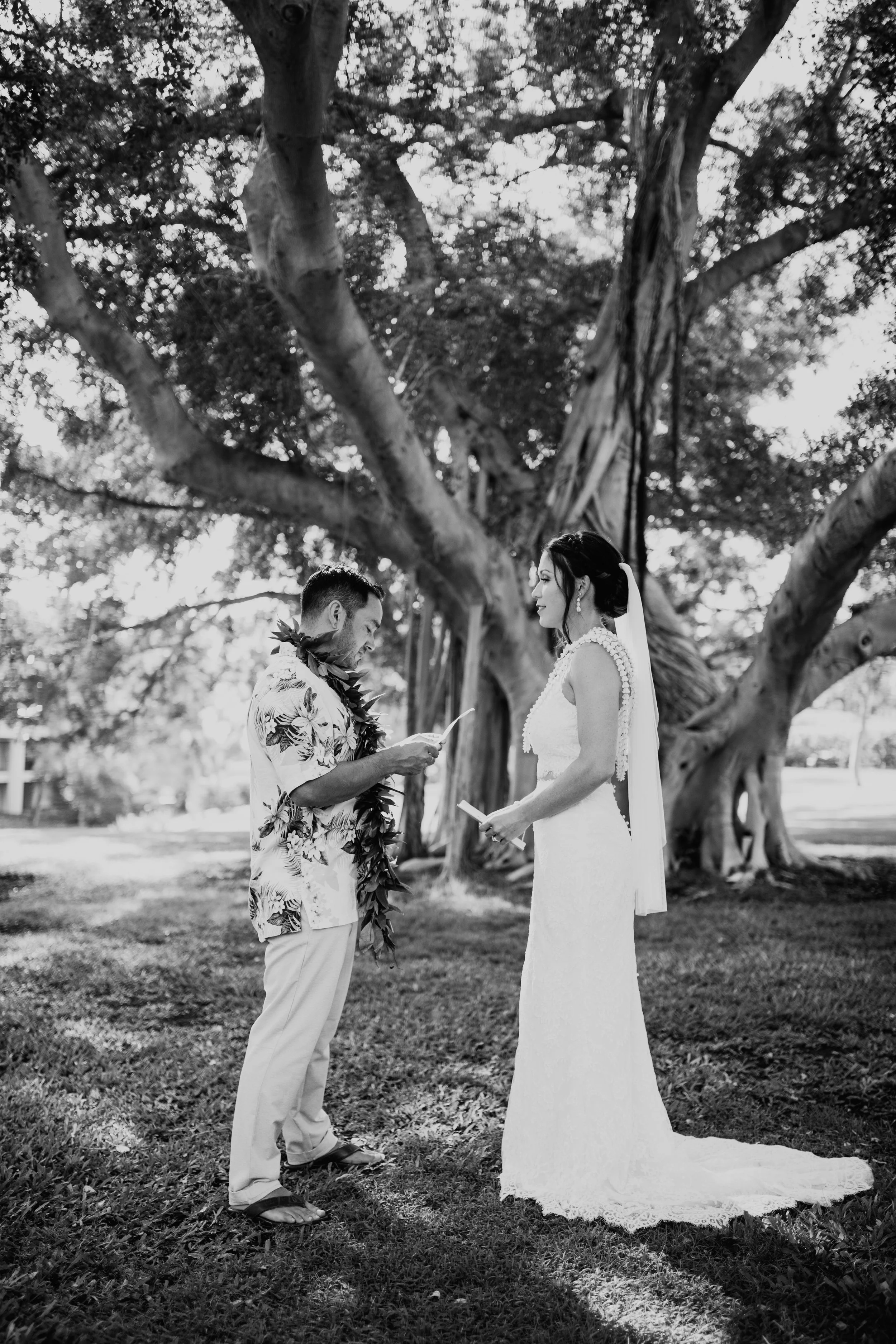 A bride and groom exchange vows outdoors under a large tree, with the groom reading from a paper and the bride holding a book, surrounded by lush greenery in black and white.