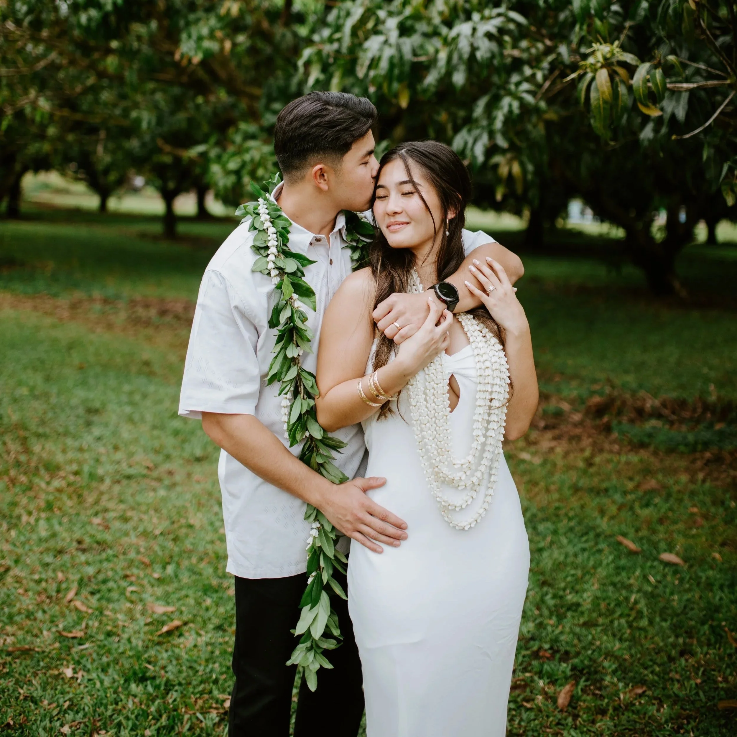 A couple in white attire sharing an affectionate moment outdoors among green trees, with the man kissing the woman's forehead and she smiling with eyes closed, wearing multiple necklaces and bracelets.