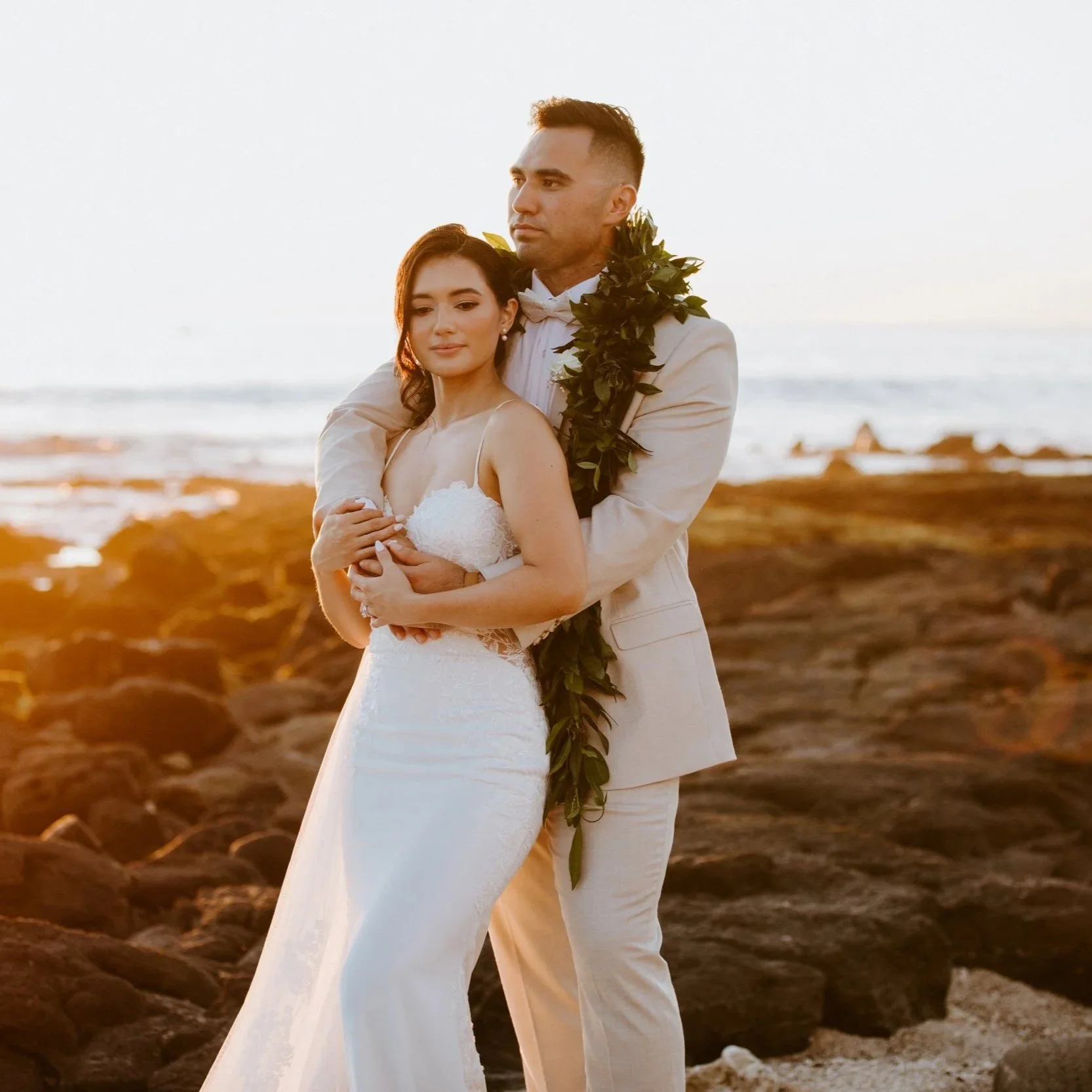 A bride and groom standing on a rocky beach at sunset, with the man wearing a cream suit and the woman in a white wedding dress, embracing each other with the ocean in the background.