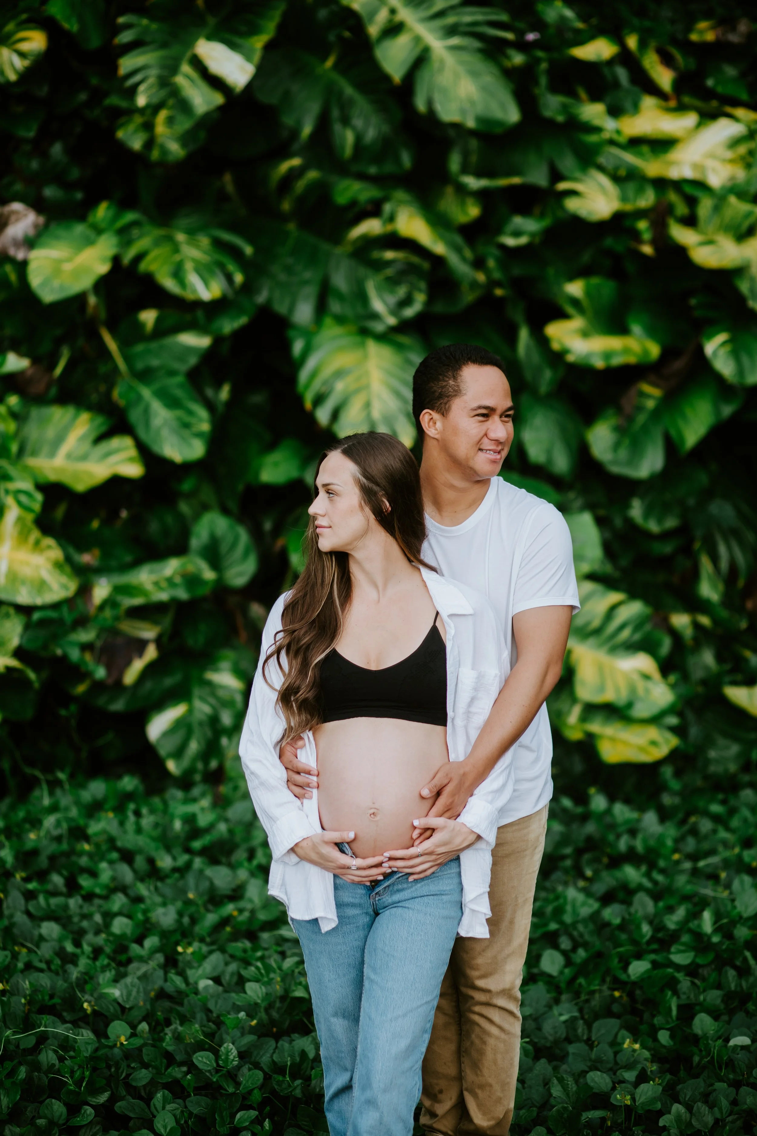 A pregnant woman and her partner standing outdoors in front of lush green foliage, with the man gently holding her belly.