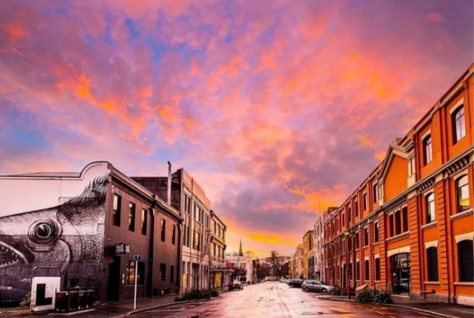 A city street at sunset with colorful orange and pink clouds and historic buildings lining both sides.