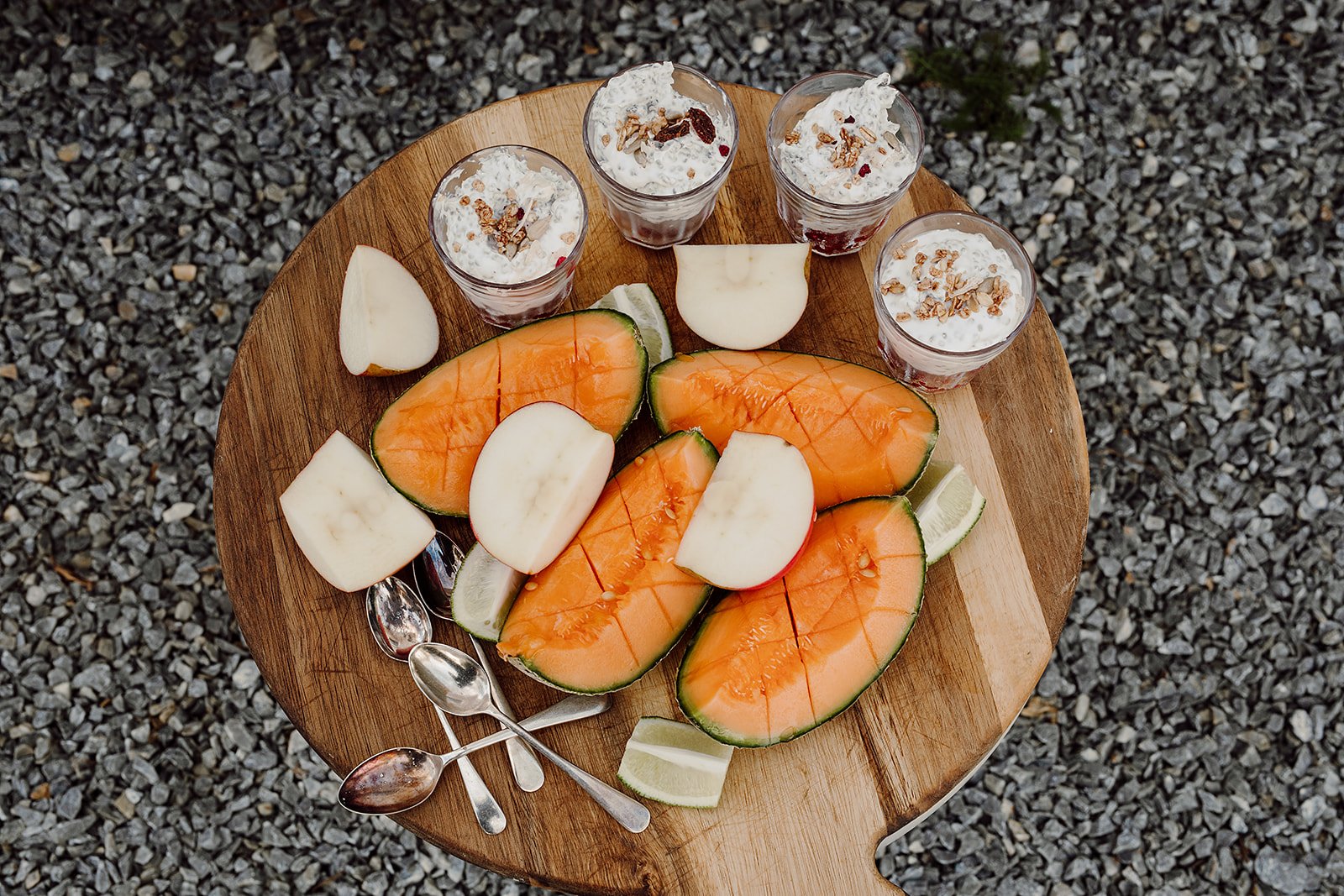 A wooden table with sliced cantaloupe, apple, and lime, along with four glasses of yogurt topped with granola, set outdoors on gravel terrain.