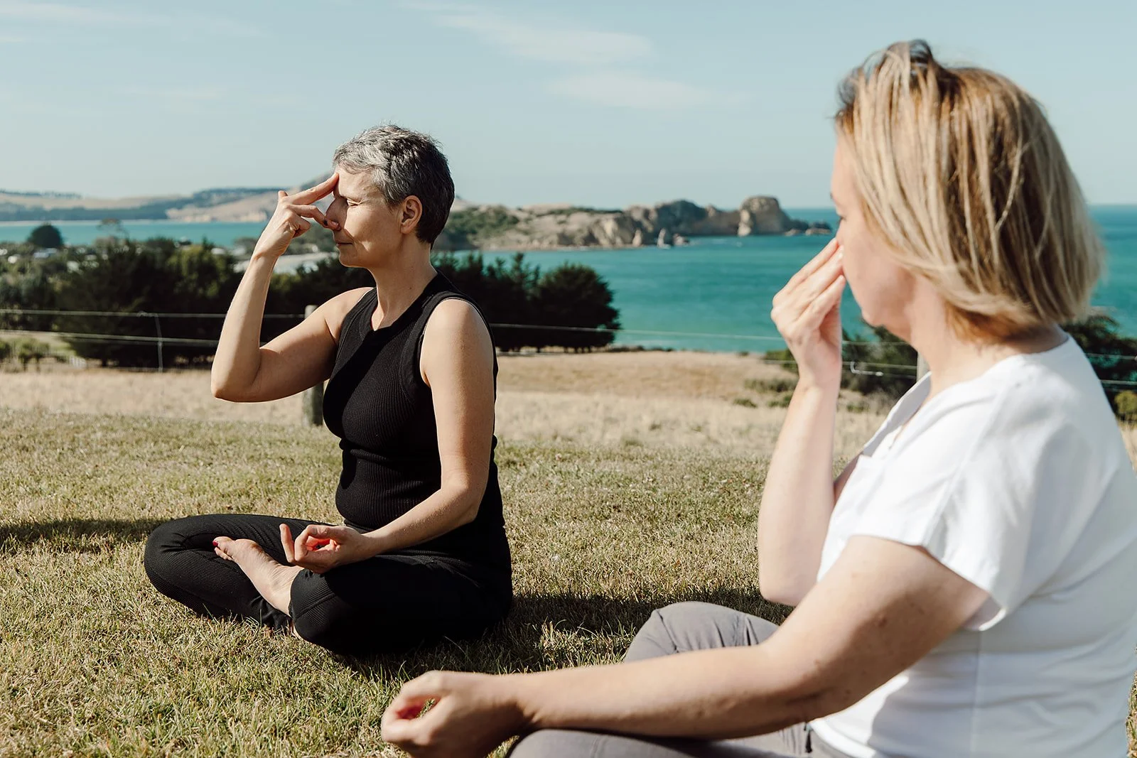 Two women sitting on grass outdoors, practicing meditation or yoga, with a scenic view of water and rocky islands in the background under a clear sky.