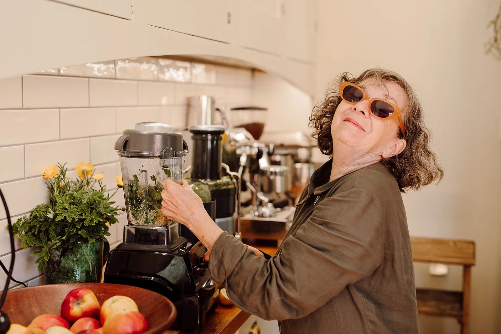 An elderly woman with curly hair wearing sunglasses is blending ingredients in a kitchen blender. She is smiling and appears to be enjoying the activity. There are apples in a bowl on the kitchen counter and a potted plant with yellow flowers nearby.