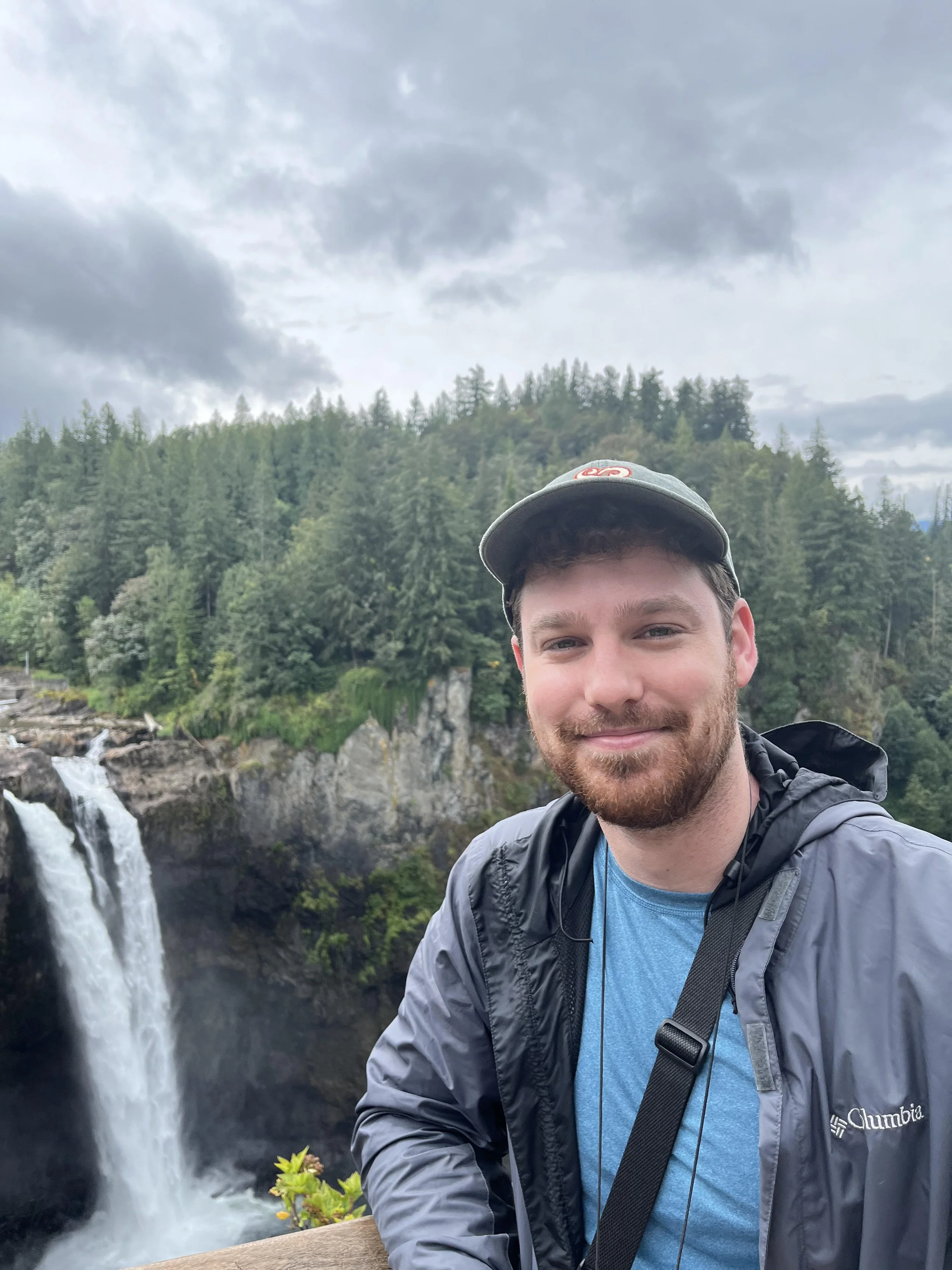 Person smiling in front of a waterfall surrounded by a forest landscape and overcast sky.