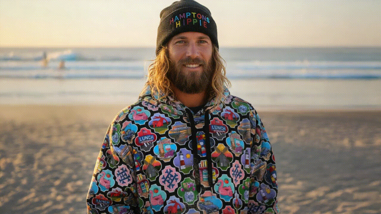 A man with long, wavy blonde hair and a beard, smiling at the camera on the beach during sunset, wearing a black beanie that says 'HAMPTON HIPPIE' and a colorful jacket with floral and peace sign patterns.