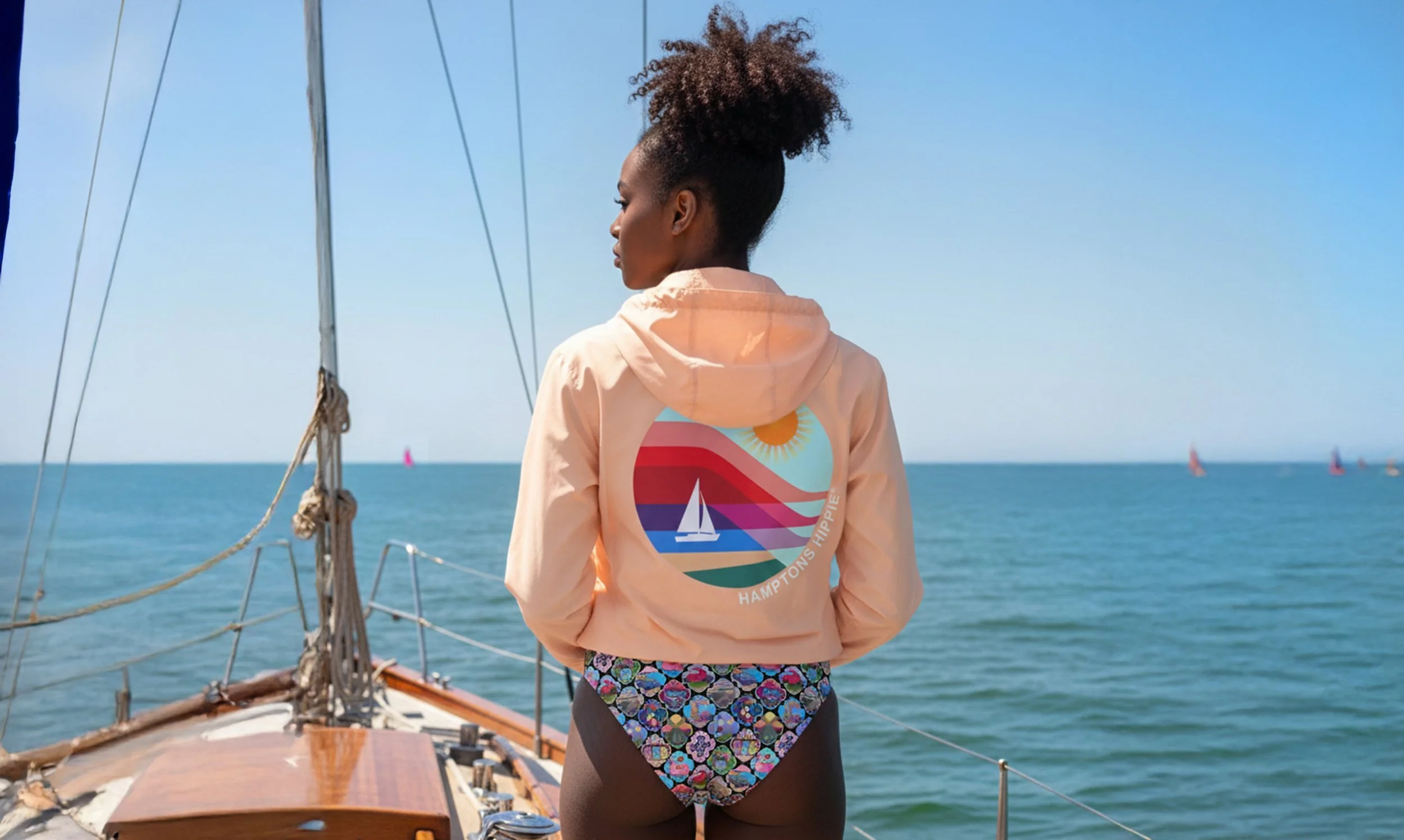 A woman on a boat looking out at sailboats with colorful sails on the water, wearing a peach hoodie with a graphic of a sailboat, sun, and stripes on the back, and floral bikini bottoms.