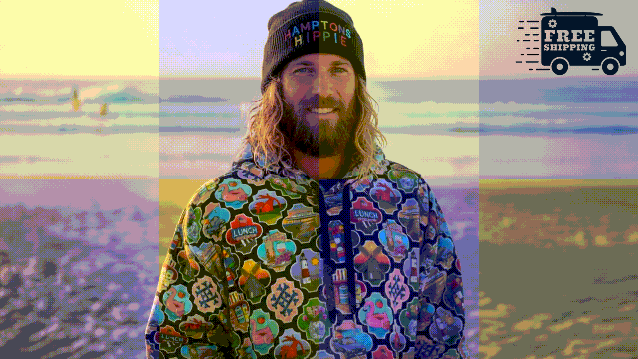 A man with long, wavy blonde hair and a beard, smiling at the camera on the beach during sunset, wearing a black beanie that says 'HAMPTON HIPPIE' and a colorful jacket with floral and peace sign patterns, plus a FREE SHIPPING visual in the right cor