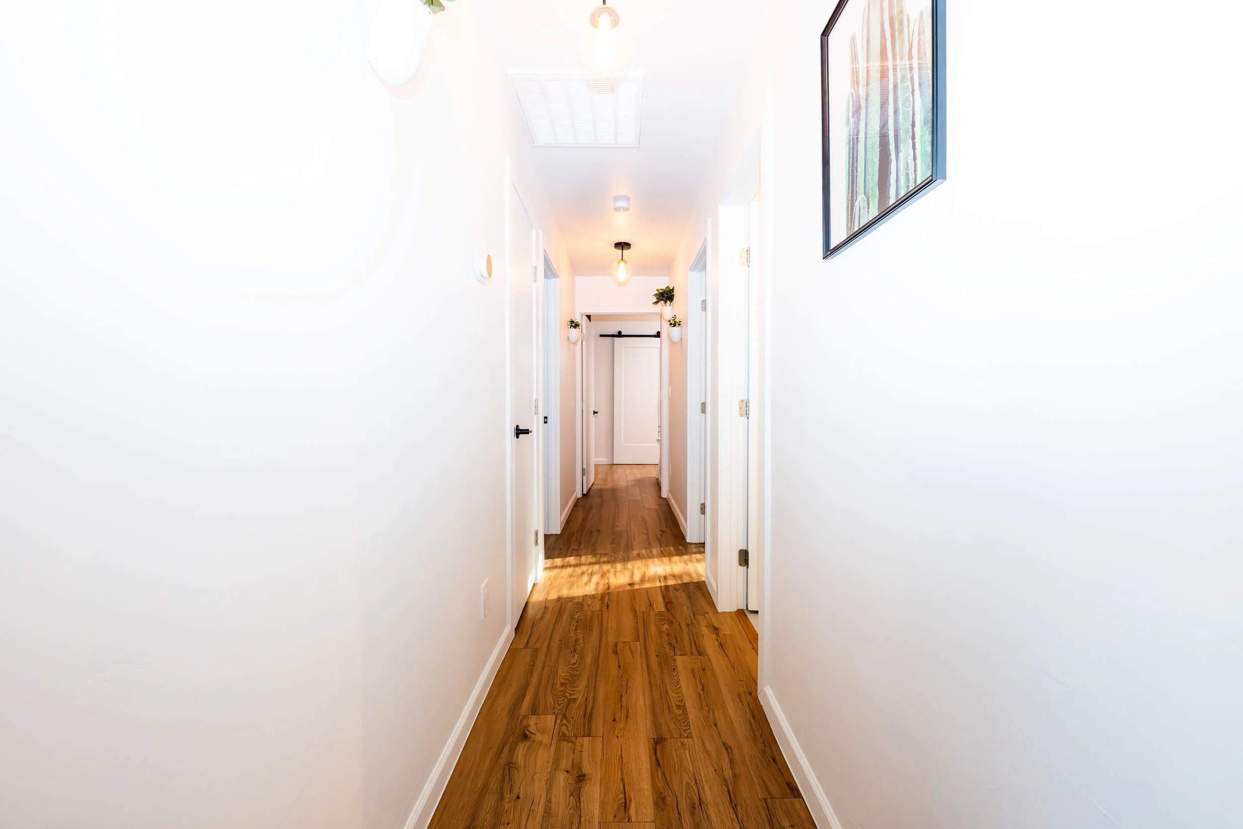 A bright, narrow hallway with white walls, wooden flooring, and ceiling lights. There are doorways on each side, with one featuring a hanging black curtain track, and a picture on the right wall.