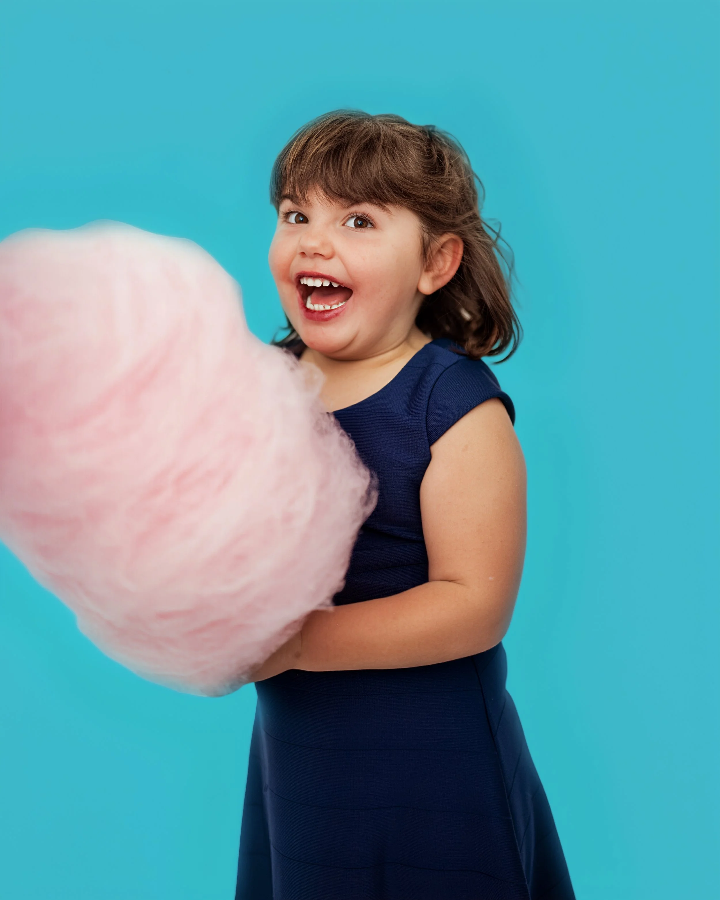 Young girl with brown hair, wearing a dark blue dress, holding pink cotton candy, smiling excitedly against a bright blue background.