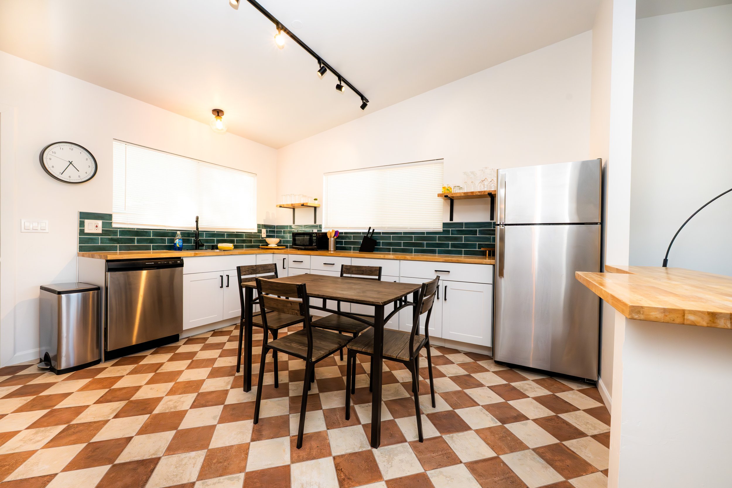 Modern kitchen with white cabinets, green backsplash, stainless steel appliances, a wooden dining table with four chairs, and checkered tile flooring.