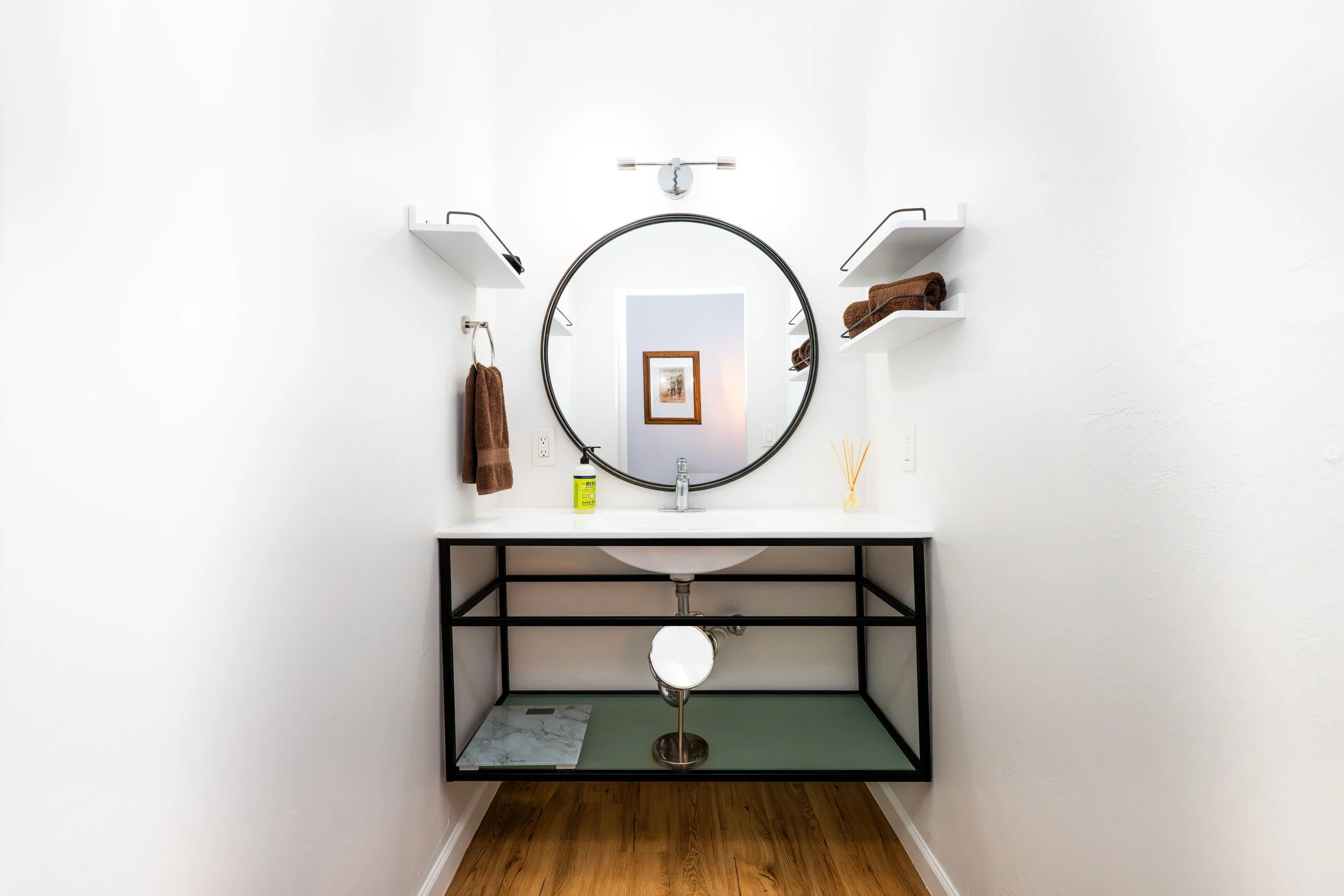 Modern bathroom vanity with oval mirror, white countertop, brown towels, and shelves on white walls.