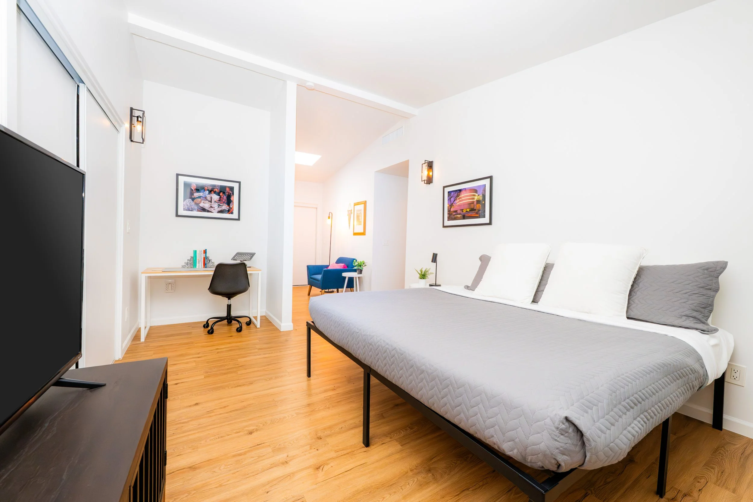 Modern studio apartment bedroom with a bed, a small desk with a chair, a living area with a sofa and lamp, hardwood floors, white walls, and framed artwork.