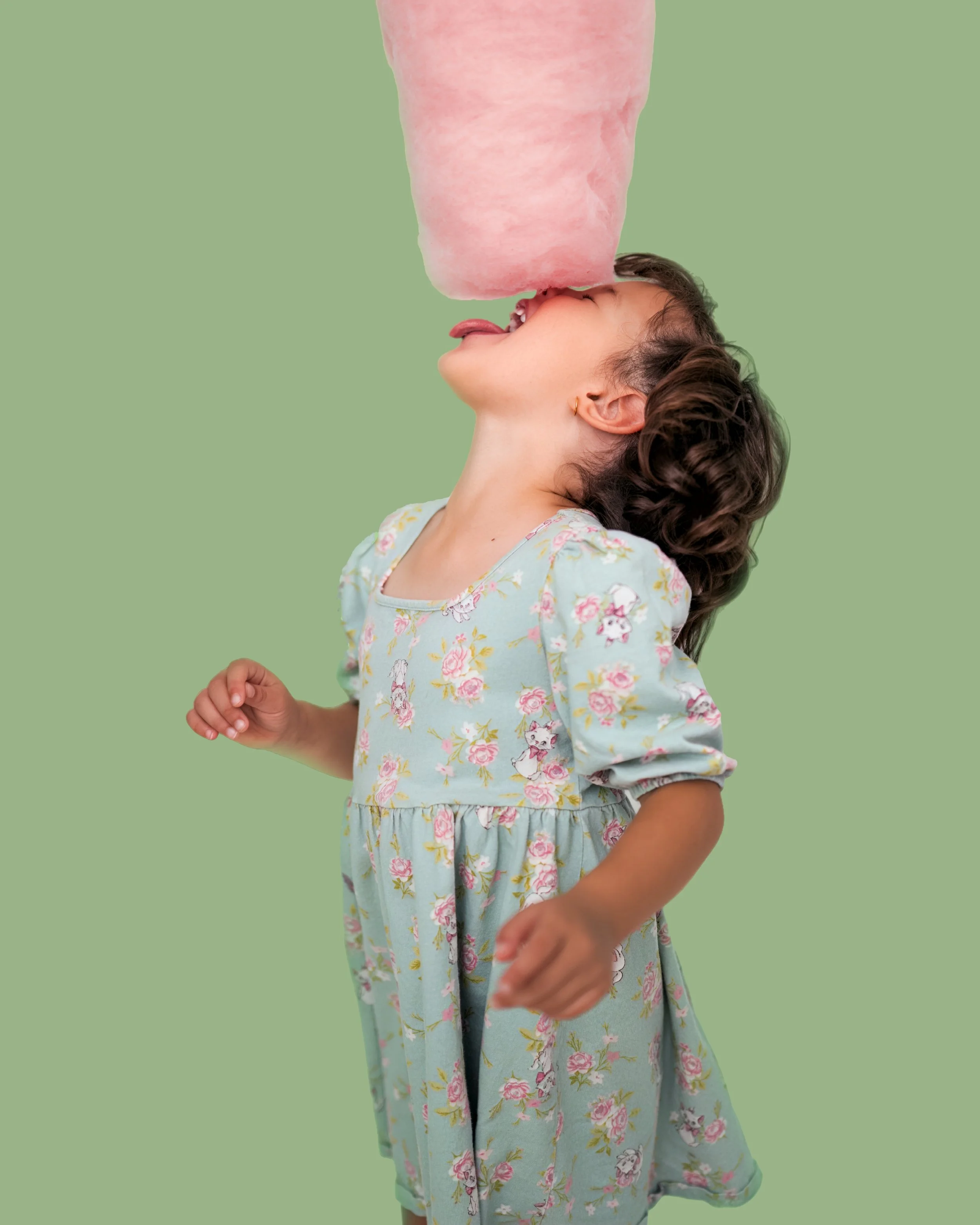 A young girl in a floral dress tilting her head back with her tongue out, licking a large pink cotton candy held above her face, against a green background.