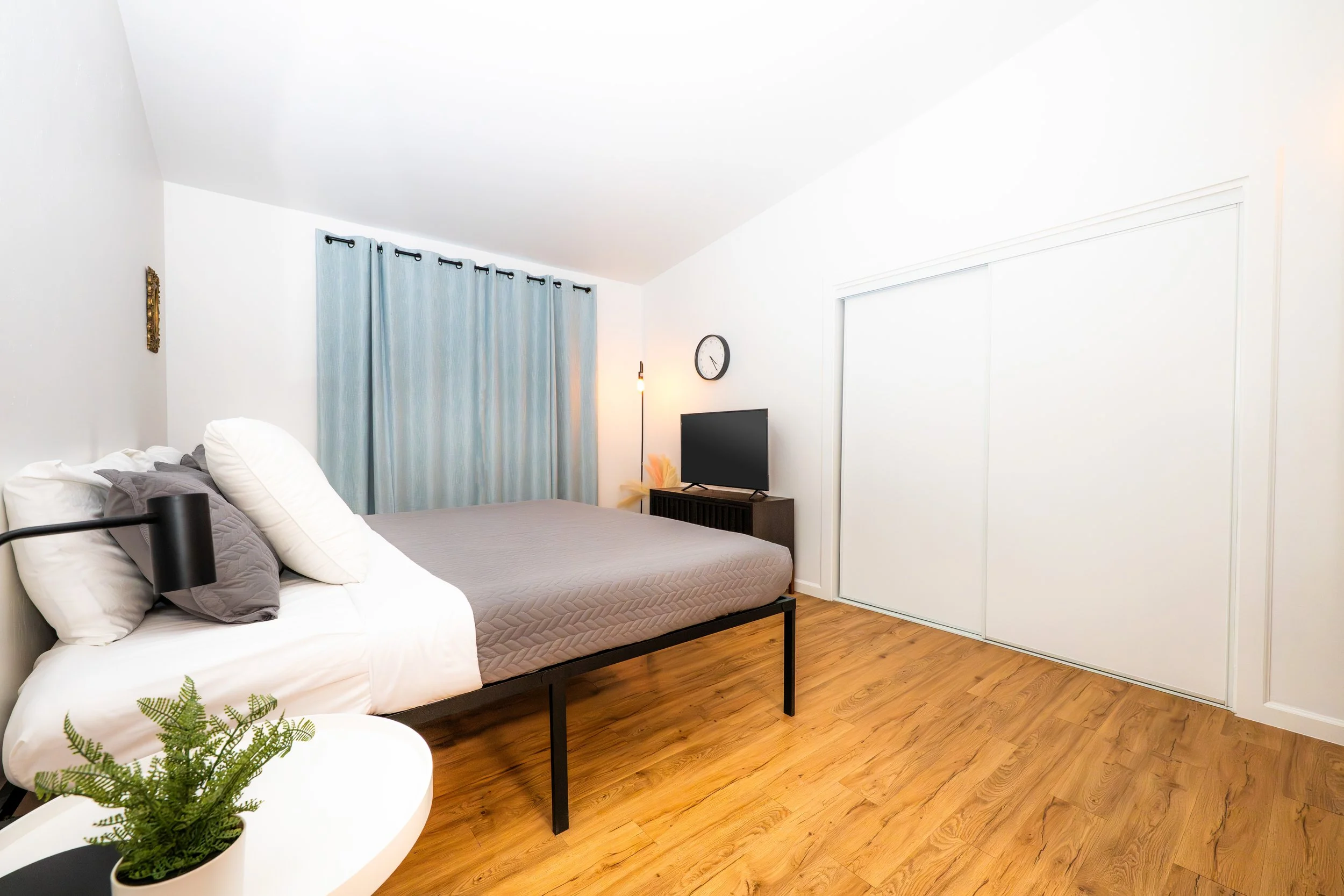 A bedroom with a bed, a window with light blue curtains, a small TV on a stand, a wall clock, a floor lamp, and a small white table with a potted plant.